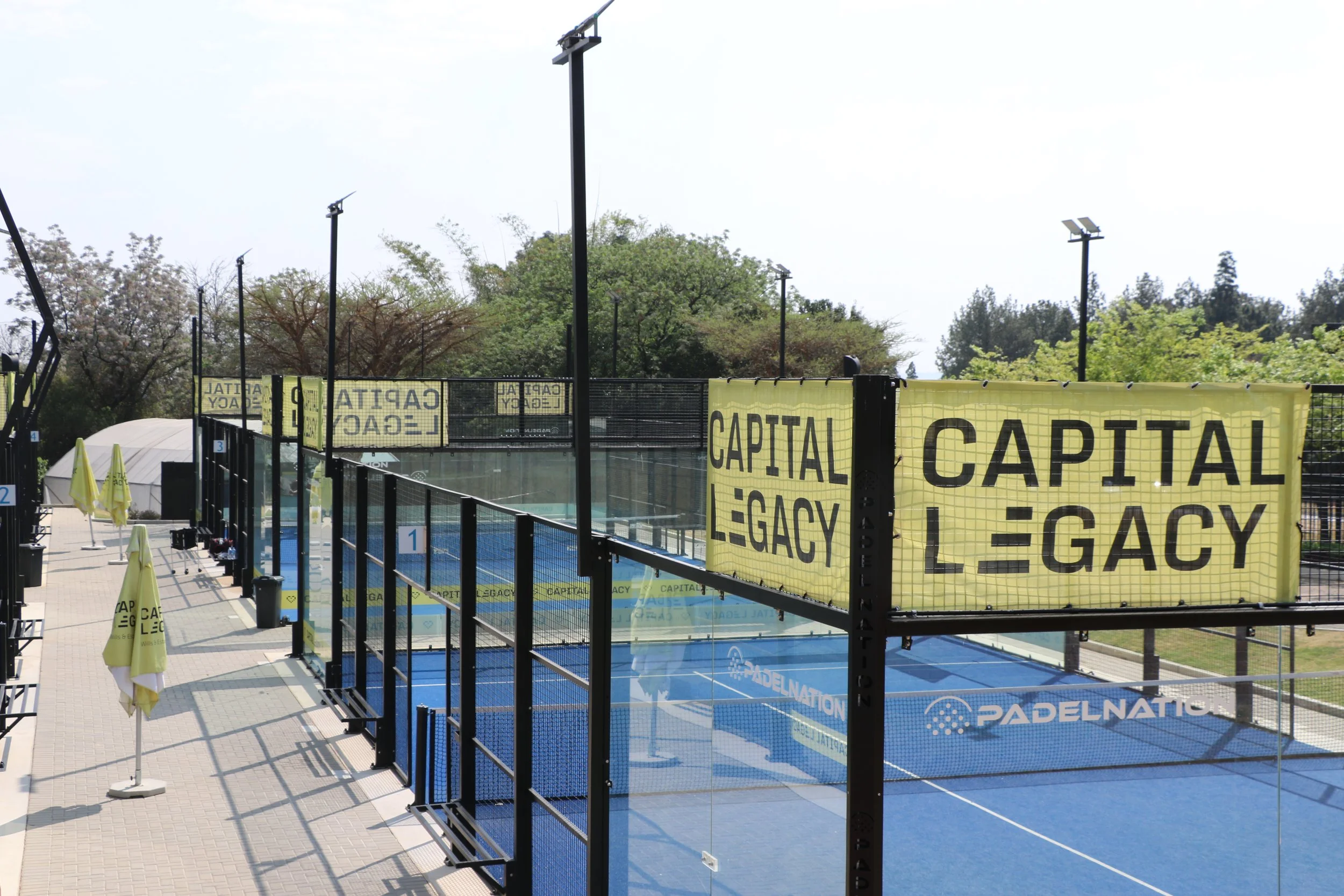 An outdoor sports complex with tennis or pickleball courts, separated by black fences and yellow banners with the words 'CAPITAL LEGACY'. There are several tall light poles, trees in the background, and umbrellas on the left side.