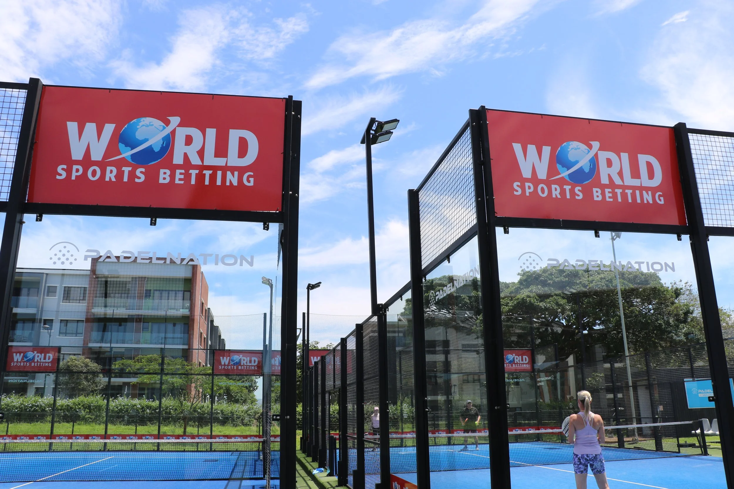 Tennis court surrounded by black fencing with parabolic LED lights and banners that read 'World Sports Betting'. The sky is mostly clear with some clouds, and there are people visible on the court and walking nearby.