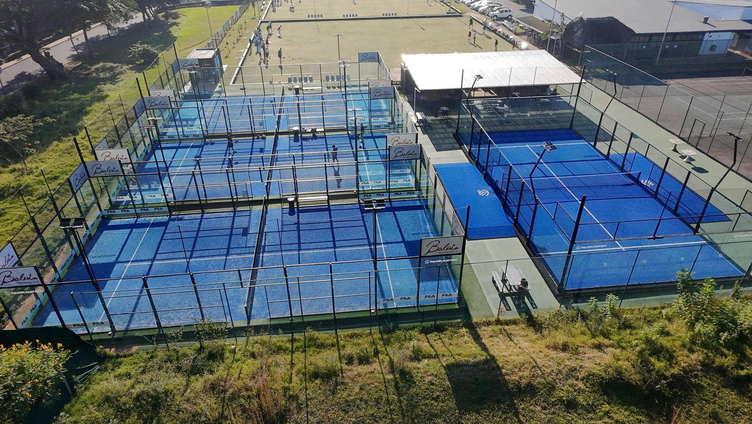 An aerial view of multiple padel tennis courts, some with players playing and others empty, surrounded by a fence and situated next to a grassy area and a parking lot.