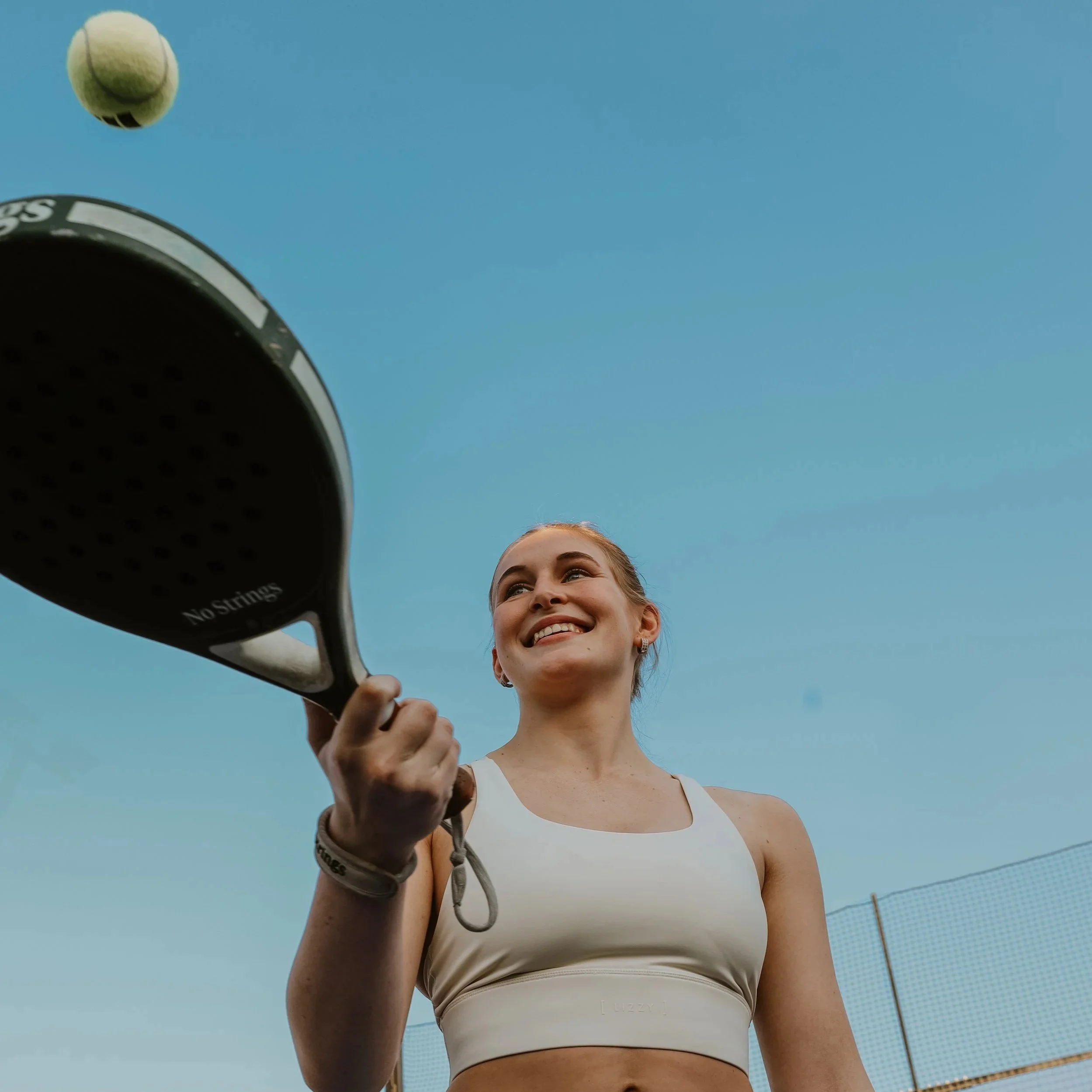 A woman bouncing a padel ball on her padel racket and smiling.