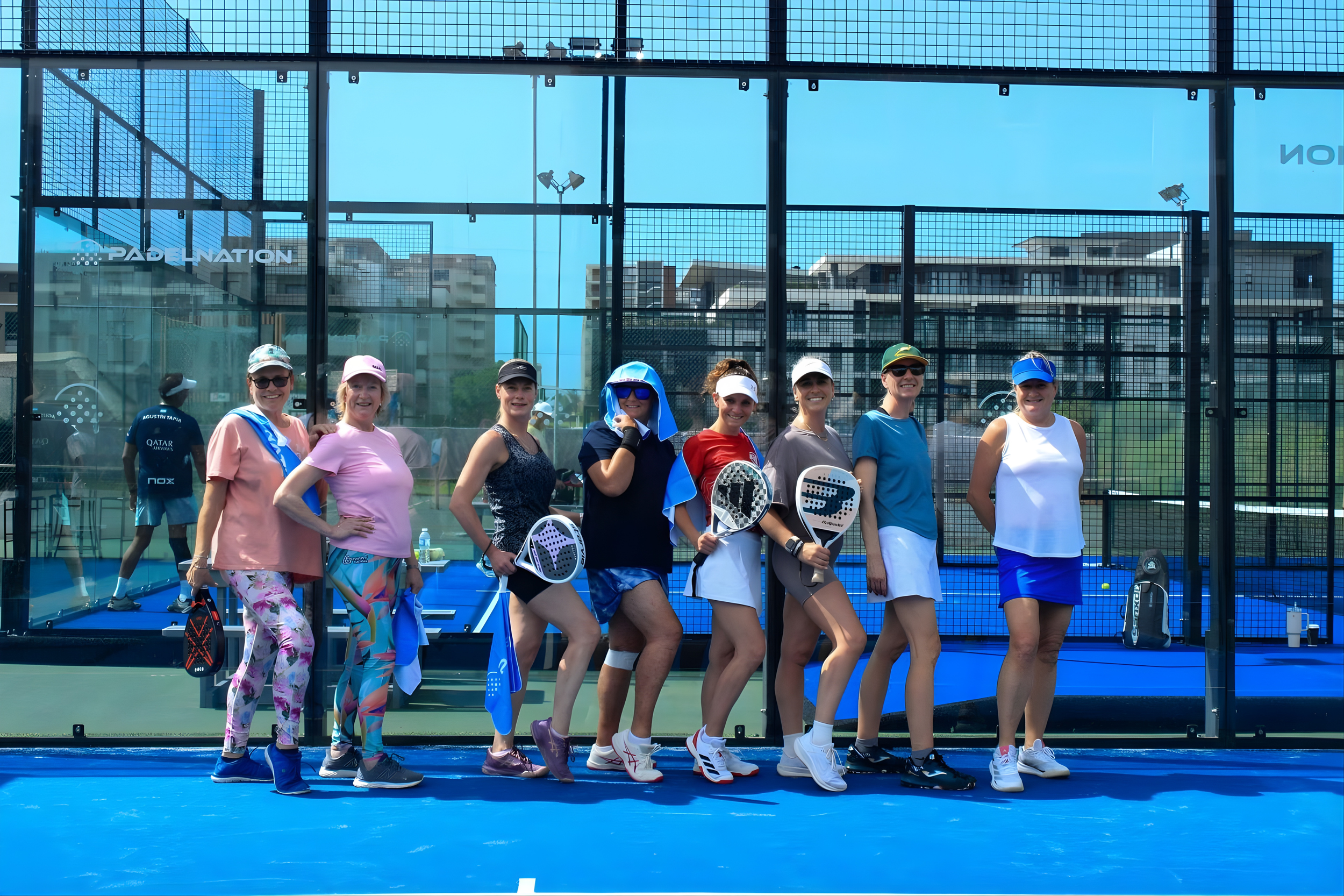 A group of women dressed in athletic clothing holding paddle tennis rackets on an outdoor court, with a glass enclosure and buildings in the background.