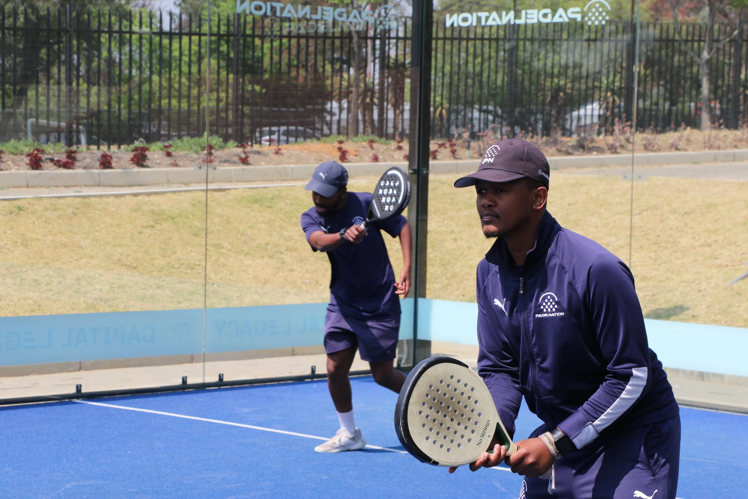 Two men in dark athletic clothing playing pickleball on an outdoor court, with one man holding a paddle and the other preparing to serve, surrounded by a black fence and trees.