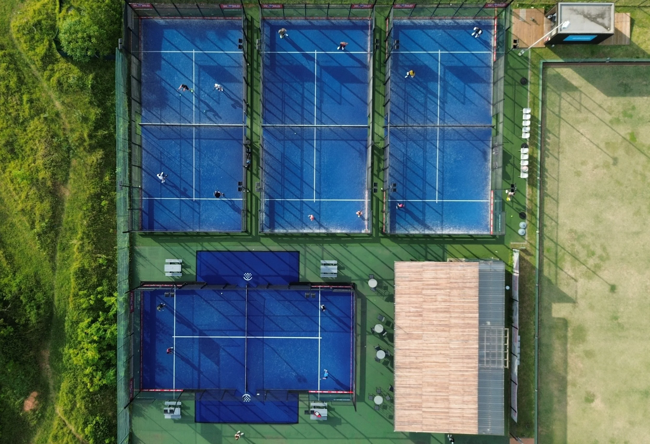 Aerial view of an outdoor tennis facility with four blue courts, some players and a surrounding green area with trees on one side.