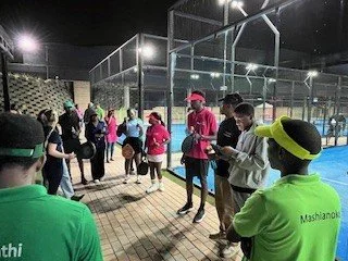 Group of people, including children, gathered outdoors near a fenced sports court at night, some wearing bright clothing and hats.
