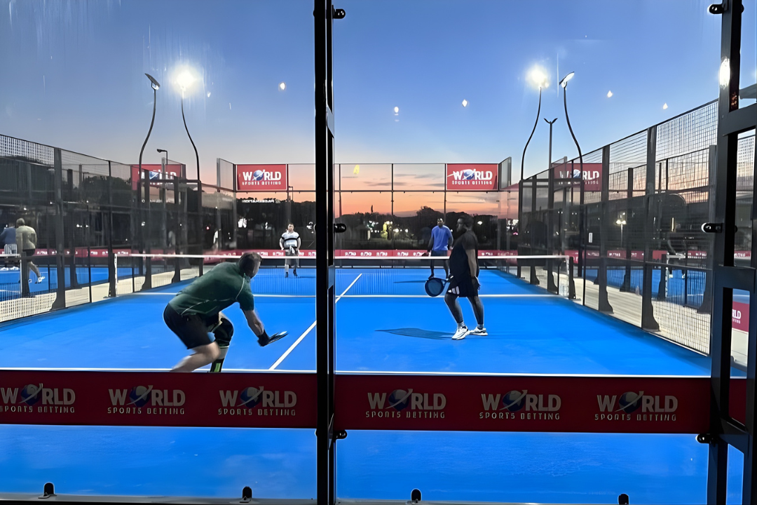 People playing paddle tennis on an outdoor court at sunset, with glass walls and net, and advertisements for 'World Sports Dressing' on the sidelines.