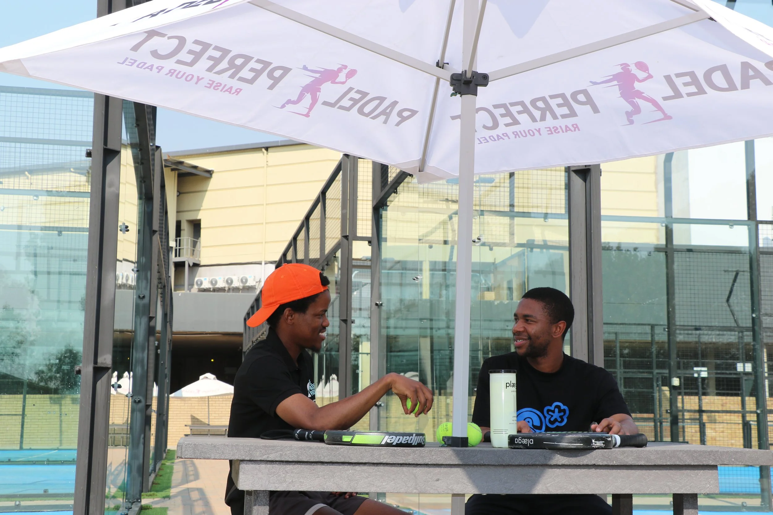 Two men sitting at an outdoor table under a white canopy with the logo 'PEACE' and a tennis player silhouette. They are shaking hands and smiling. The table has tennis equipment like a racket, tennis balls, and water bottles.