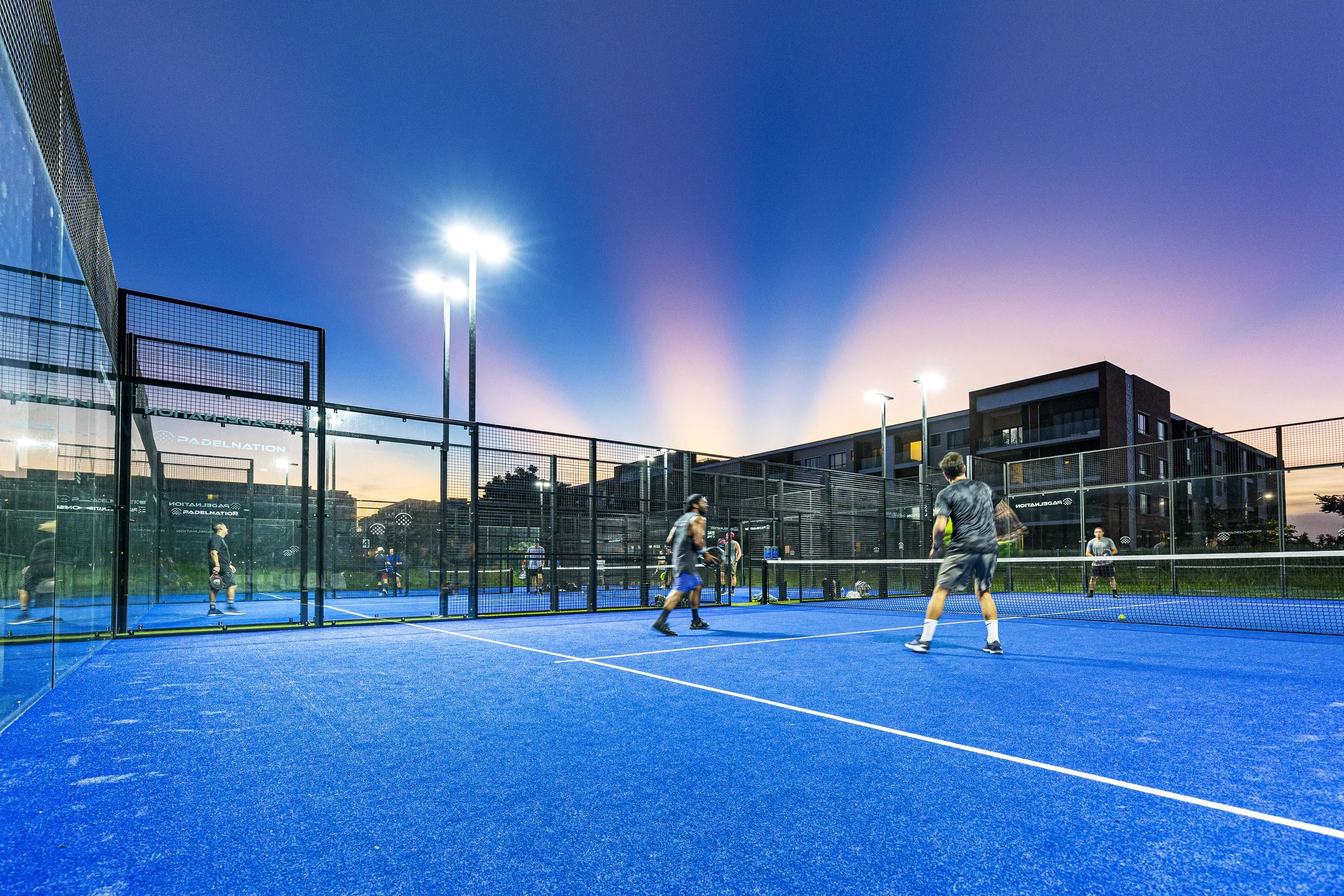 A group of people playing pickleball on an outdoor blue court during sunset, surrounded by fences and modern residential buildings.