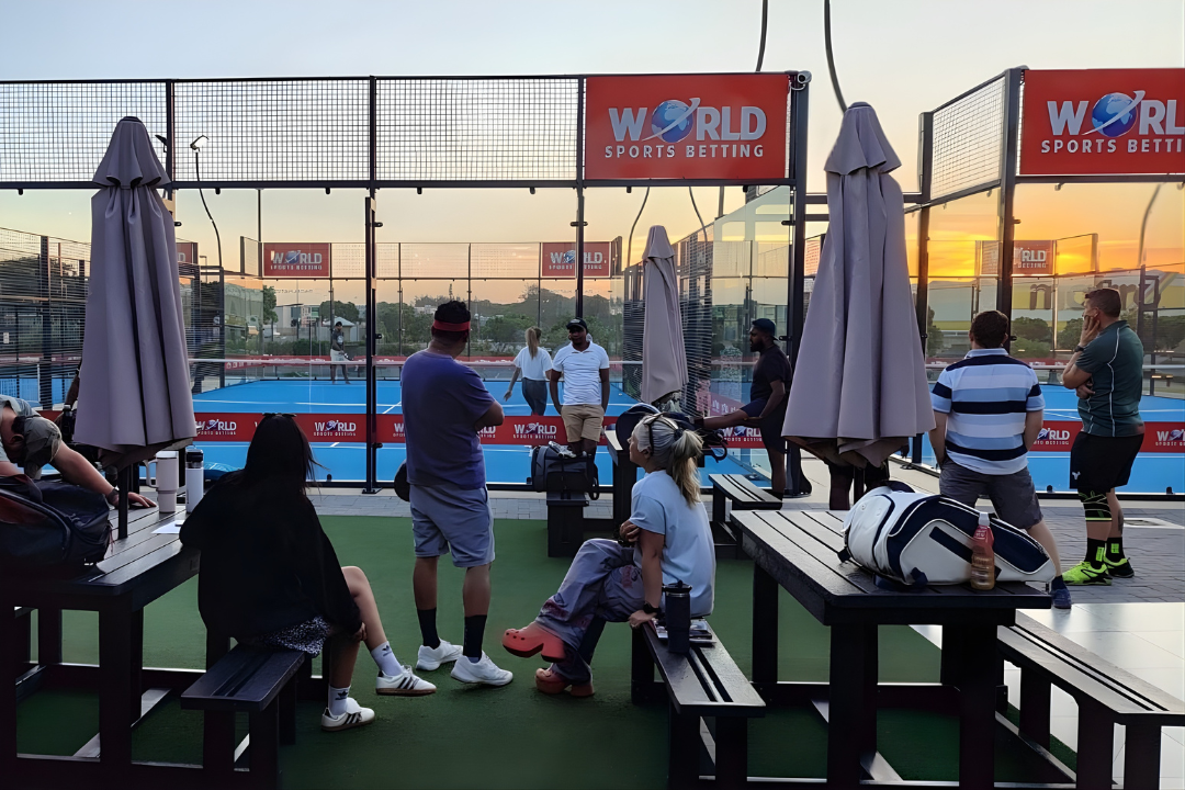 People watching a pickleball game at outdoor courts during sunset, with umbrellas and seating area.