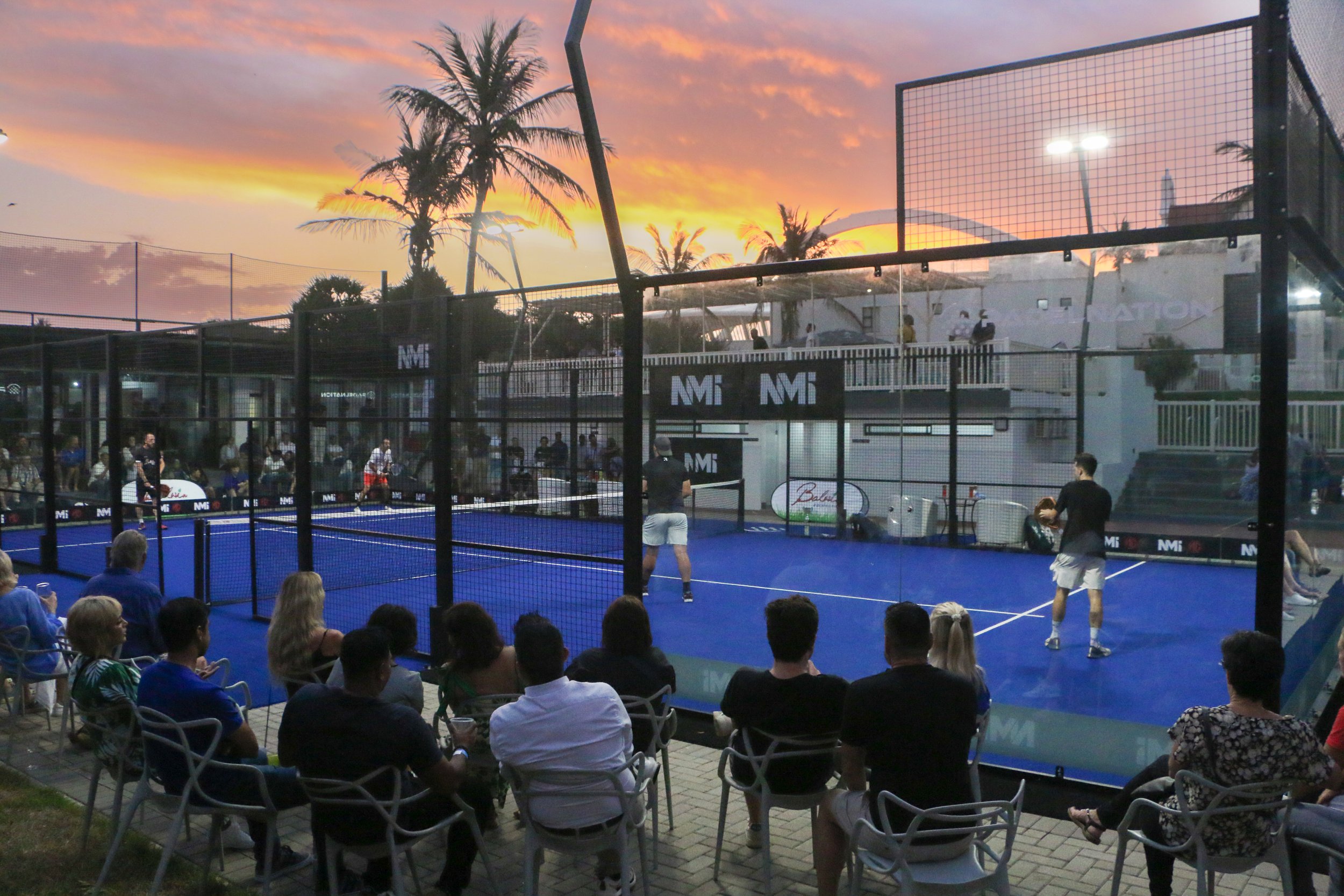 People playing pickleball on an outdoor court during sunset, with spectators seated nearby and palm trees in the background.