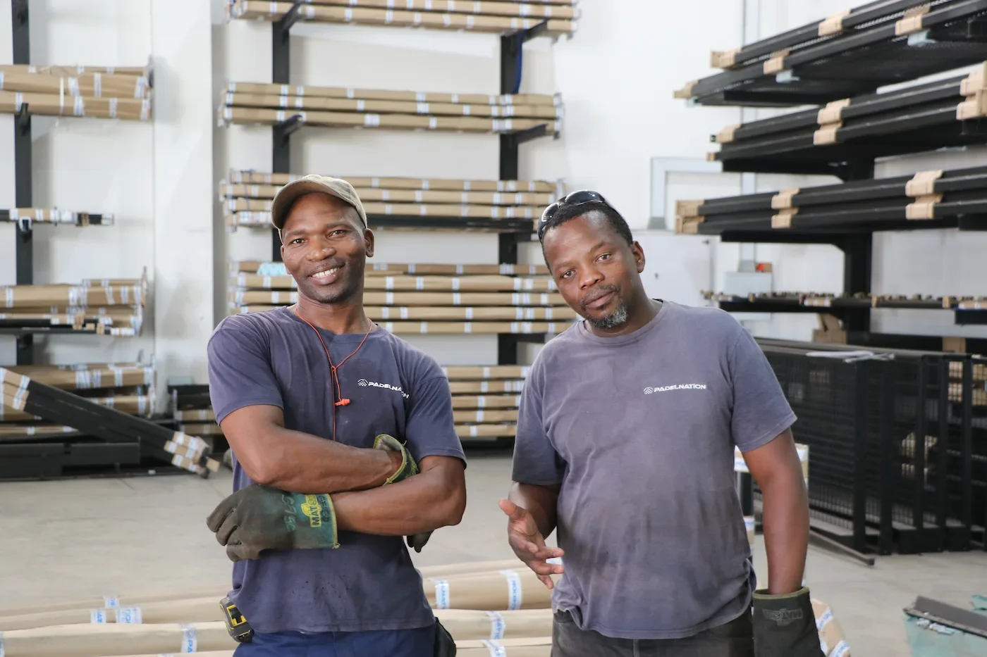 Two smiling men in casual work clothes and gloves standing inside PadelNation's padel court factory, based in Tongaat.