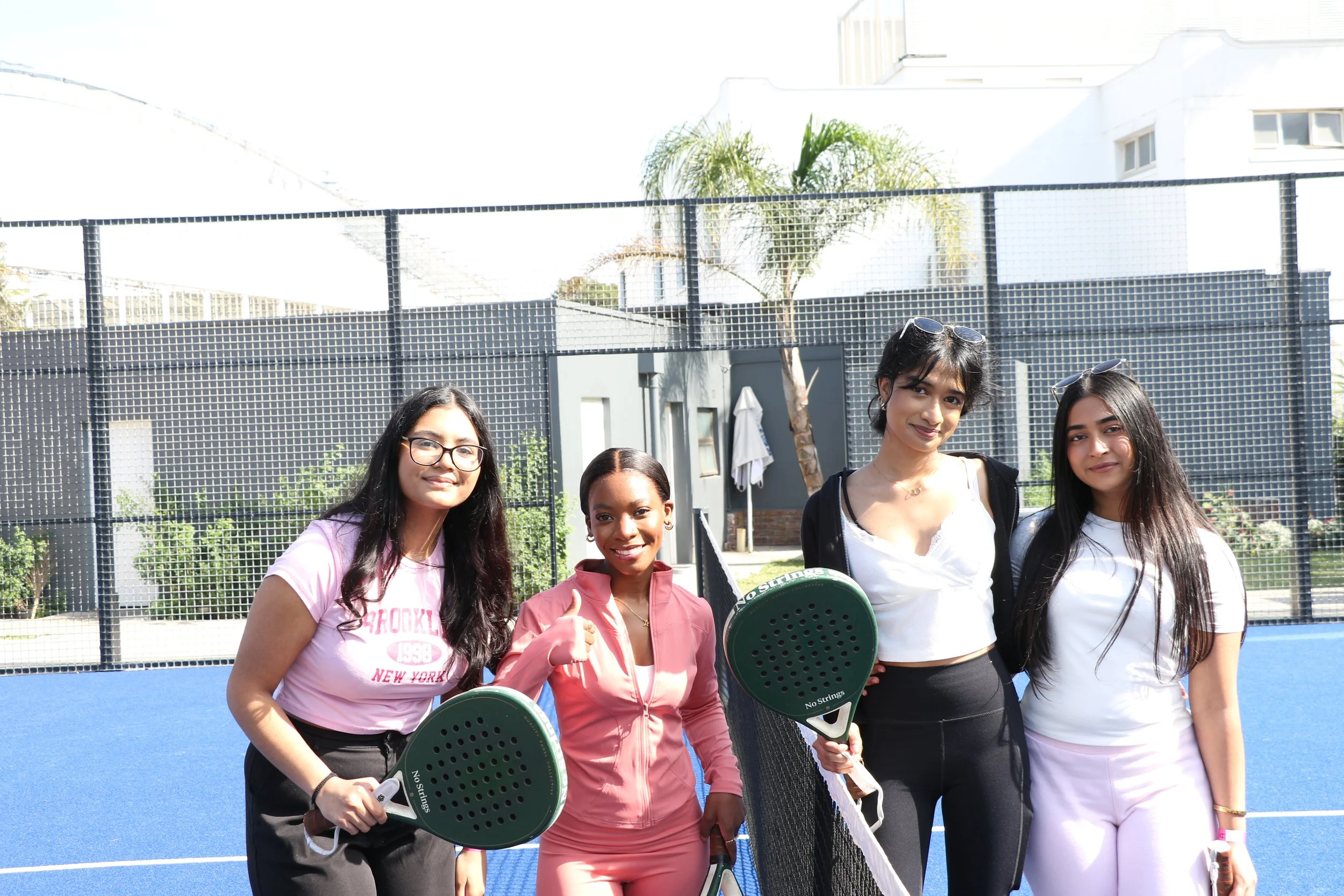 Four young women standing on a tennis court, holding pickleball paddles, smiling at the camera.