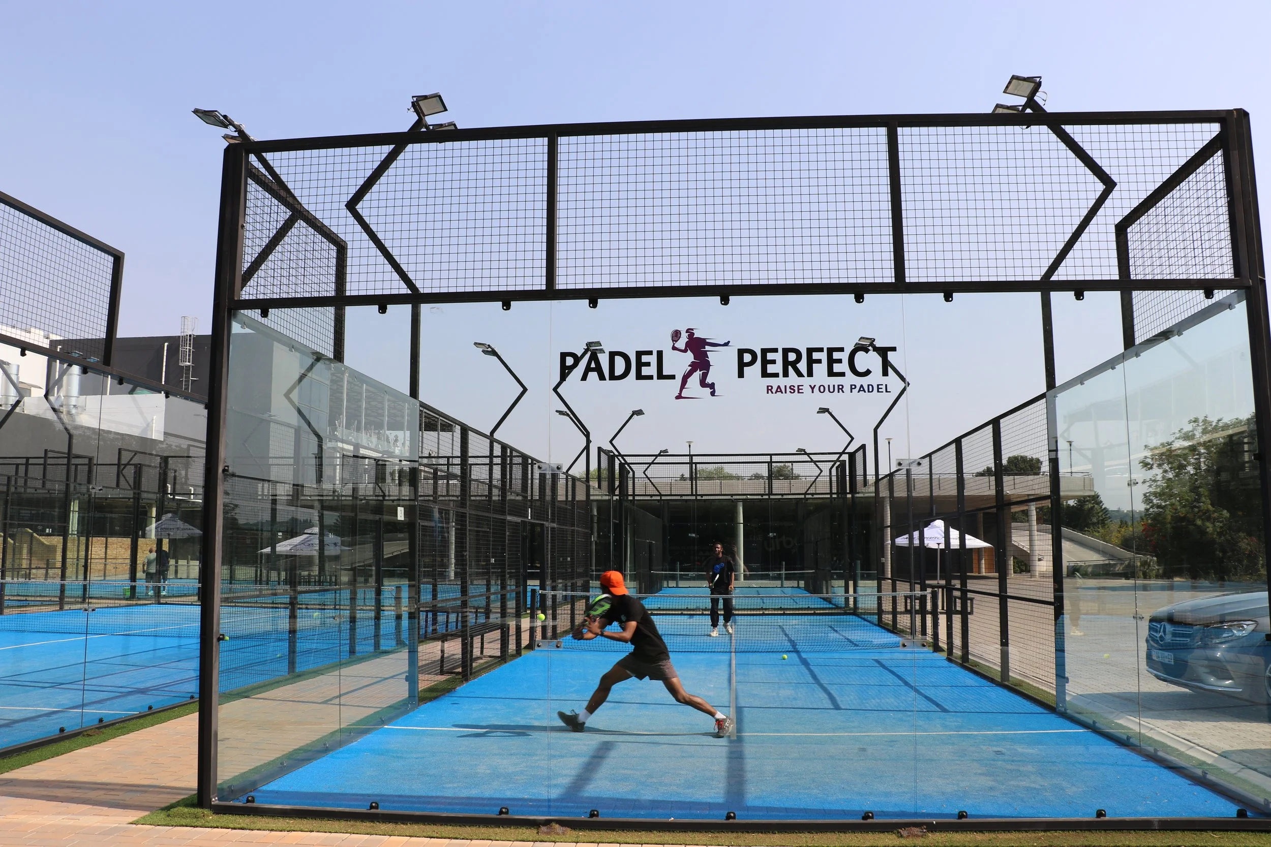 An outdoor padel tennis court with blue playing surface surrounded by glass walls and fencing. There are players on the court hitting a ball, and a sign overhead that reads 'Padel Perfect Raise Your Padel.'
