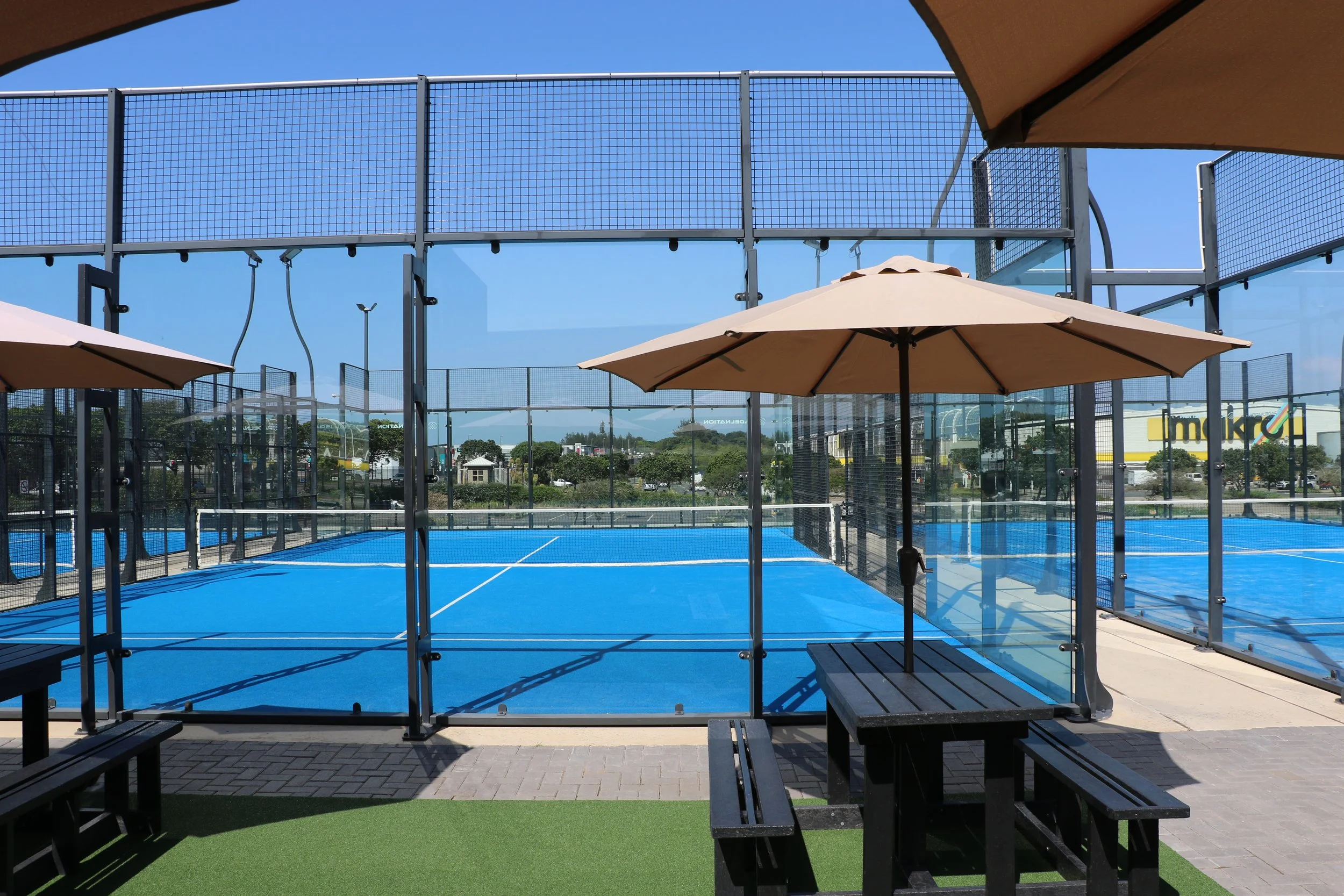 An outdoor tennis court with blue surface, surrounded by glass walls and fencing, with large umbrellas providing shade, and trees visible in the background.