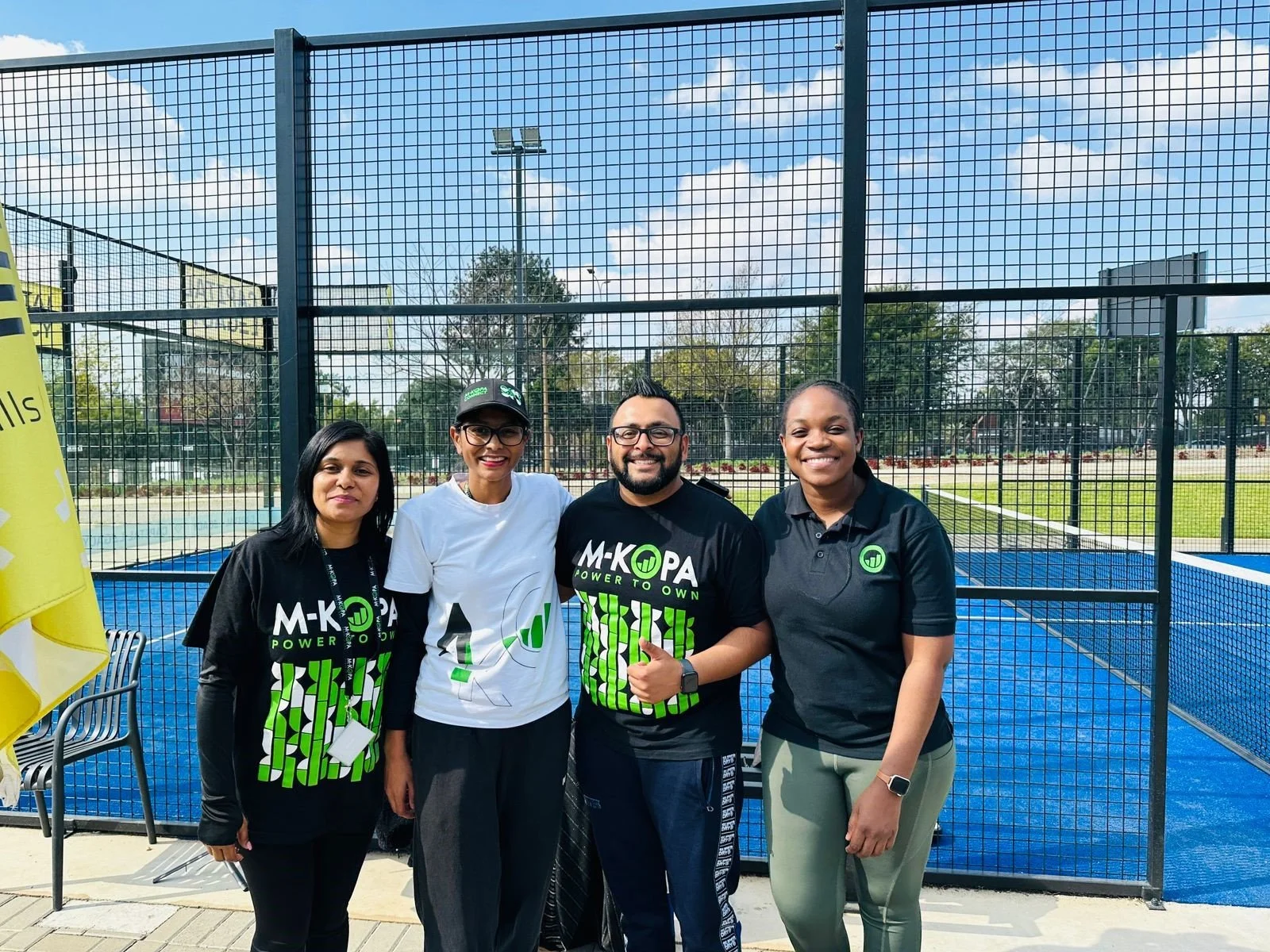 Four people standing in front of a fenced tennis court outdoors, smiling at the camera. Three women and one man, with some wearing black and green 'M-KOPA' shirts, and one woman in a white shirt and glasses.