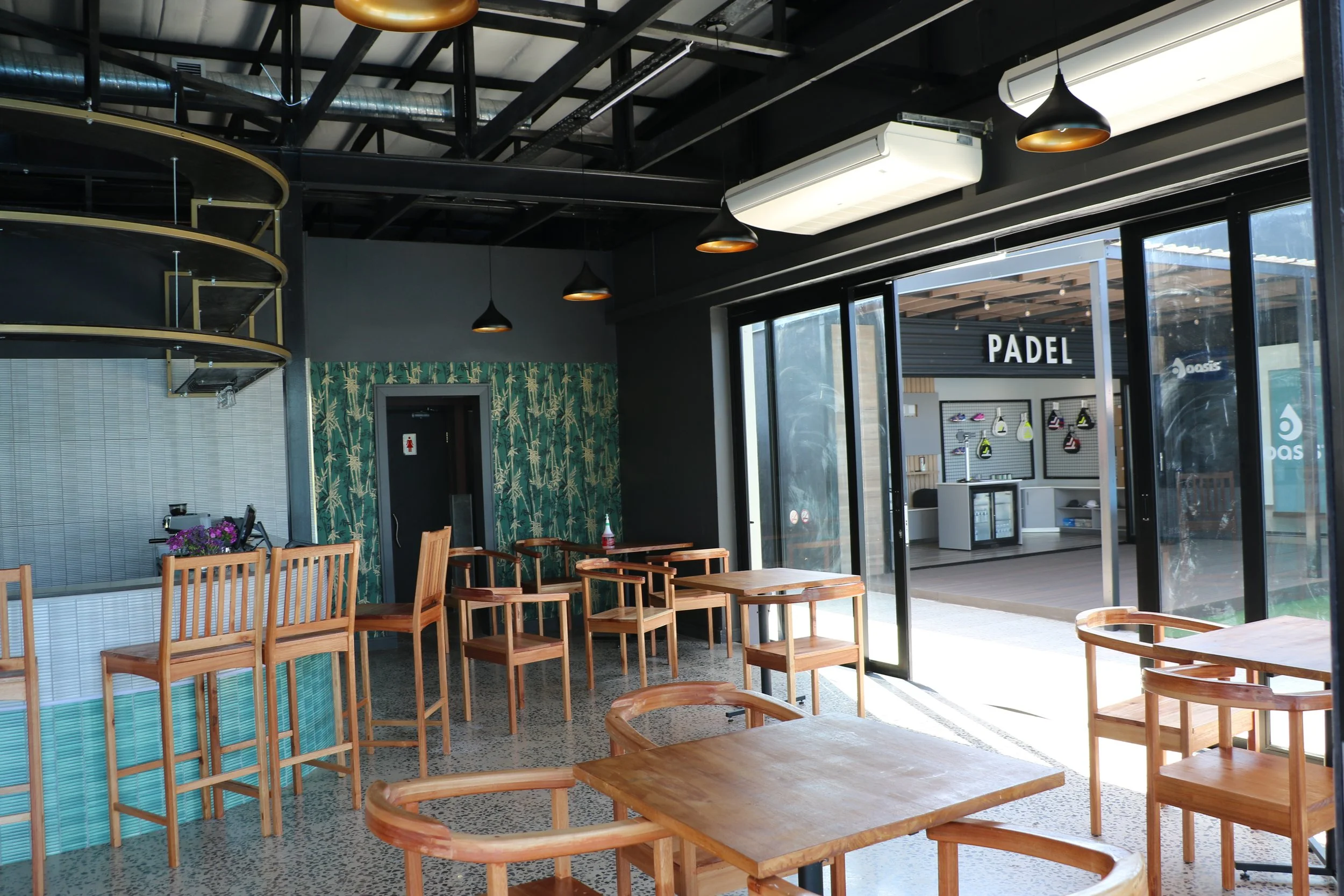 Interior of a modern cafe with wooden tables and chairs, large glass windows, and a sign that says 'PADel' outside.