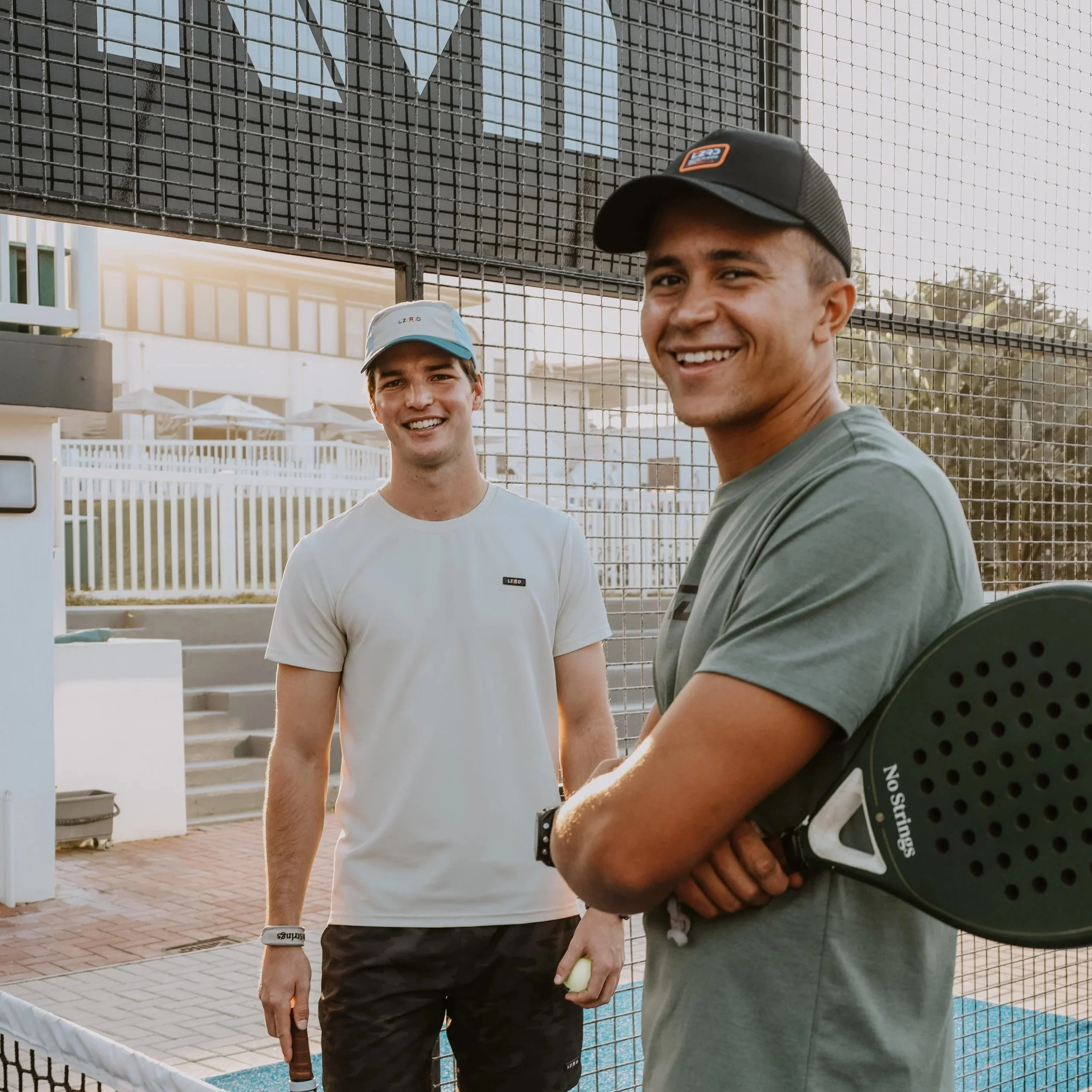 Two young men on a padel court, smiling at the camera, holding padel rackets.