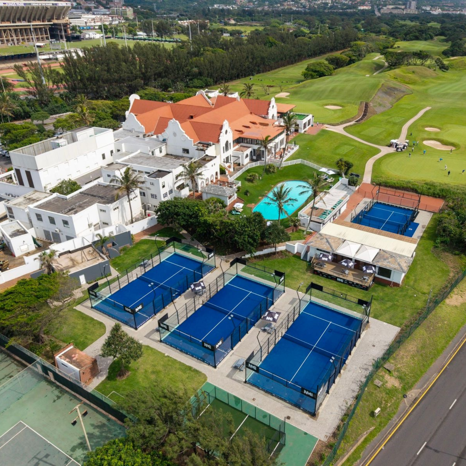 Aerial view of a sports complex featuring multiple tennis courts and a swimming pool, with a large white building and a golf course in the background.