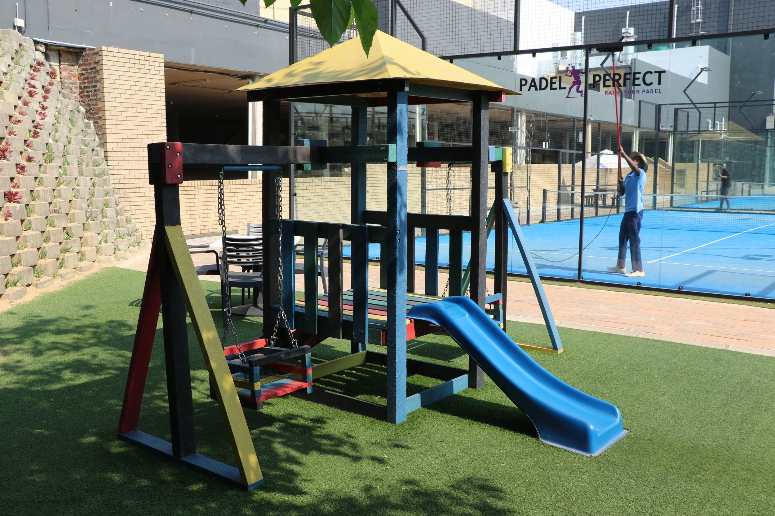 Colorful playground set with slide and swings, situated on artificial grass. In the background, a person is playing paddle tennis on a court with glass walls, and there are chairs stacked near the playground.