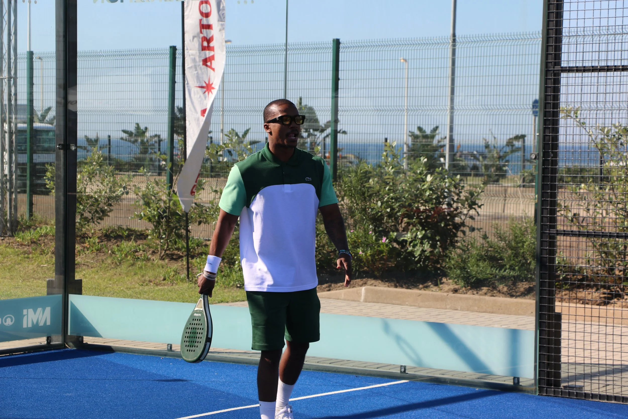 A man holding a tennis racket on a tennis court with a glass wall and a banner in the background.
