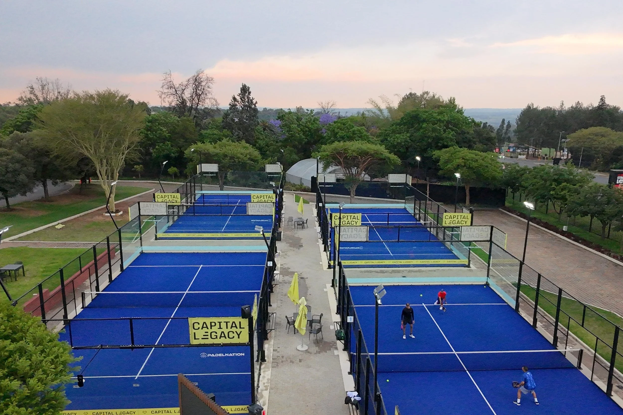 Multiple outdoor padel tennis courts with blue surfaces, yellow banners reading 'CAPITAL LEGACY,' and players practicing. Surrounding trees and a cloudy sky in the background.