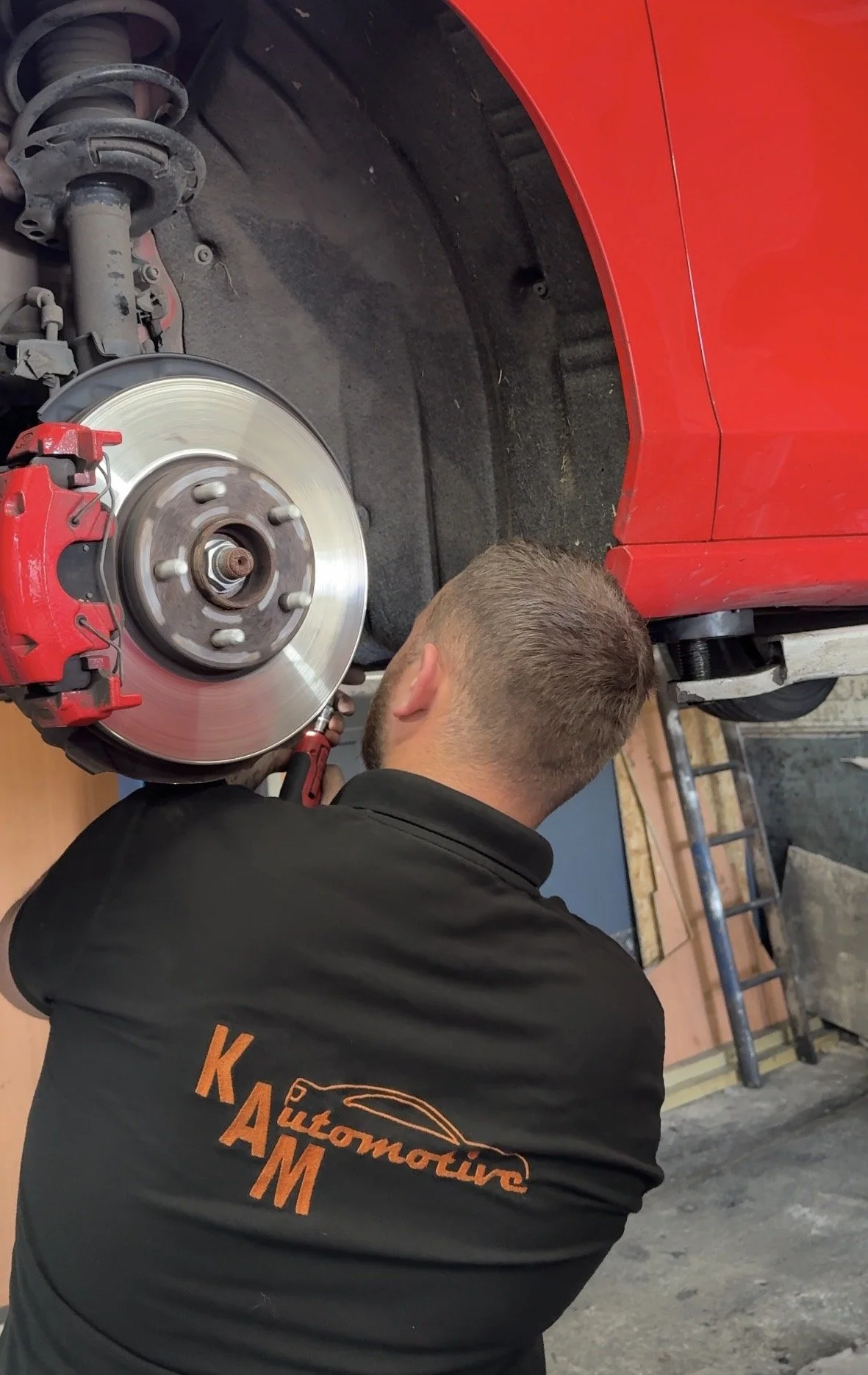 A mechanic working on a red car, removing or installing the brake disc and caliper. The mechanic is wearing a black shirt with orange lettering.