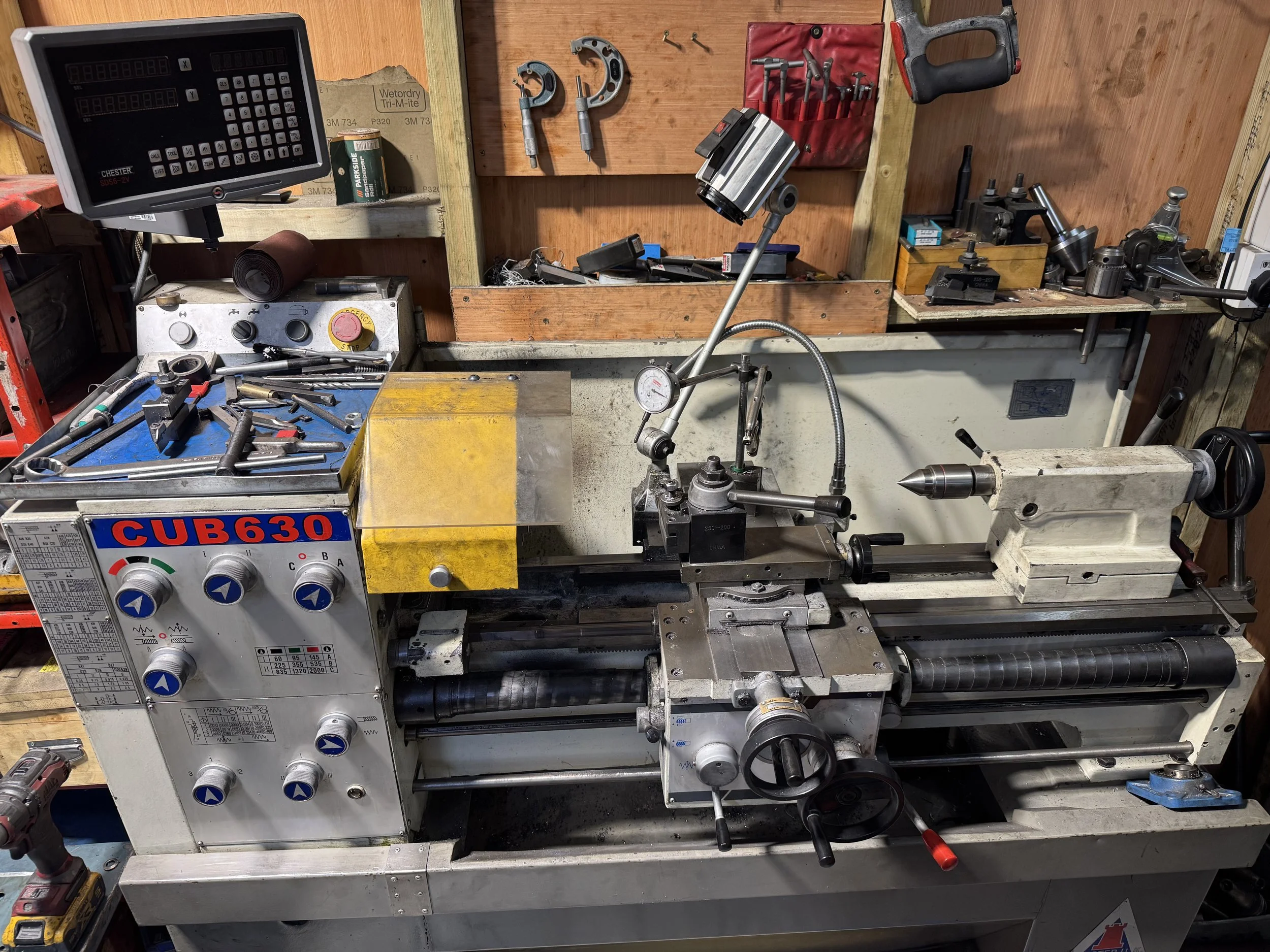 A workbench in a workshop with machining tools, including a lathe, drill bits, clamps, a power drill, and other equipment.