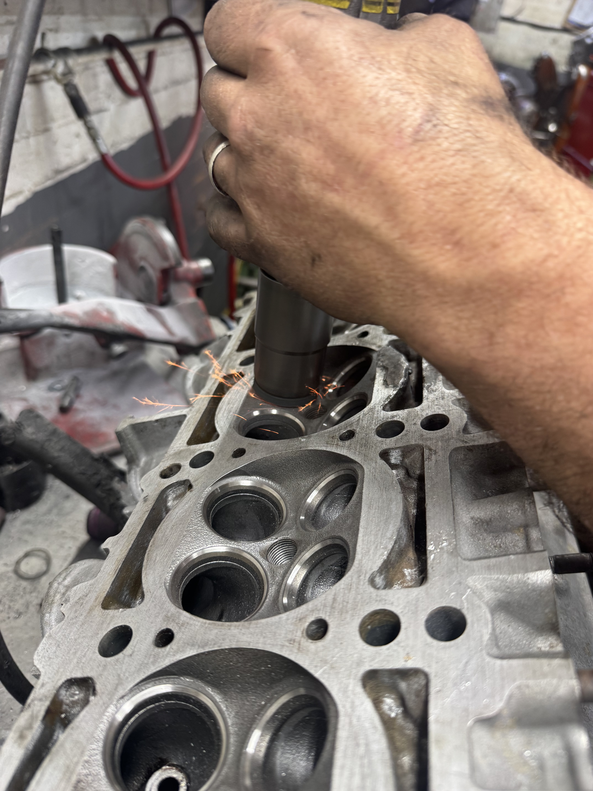 Close-up of a mechanic's hand working with a power tool on a metal engine block with multiple cylinders, sparks flying, and various tools in the background.