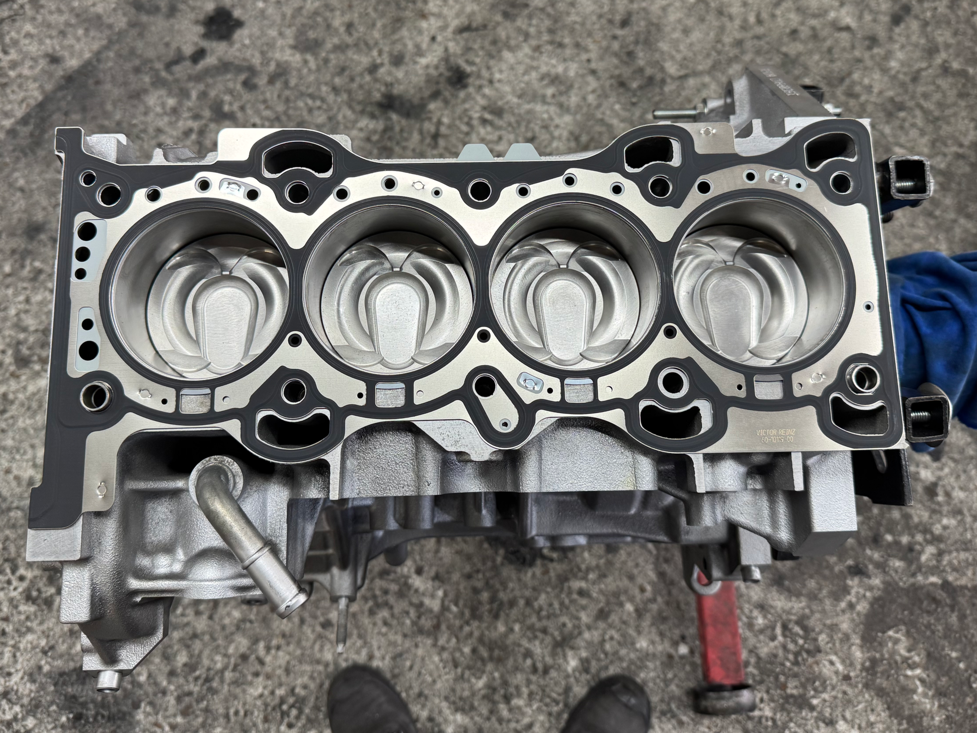 Close-up of a silver automotive engine cylinder head with three large circular combustion chambers and various drilled holes for valves and spark plugs, placed on a grey textured surface.