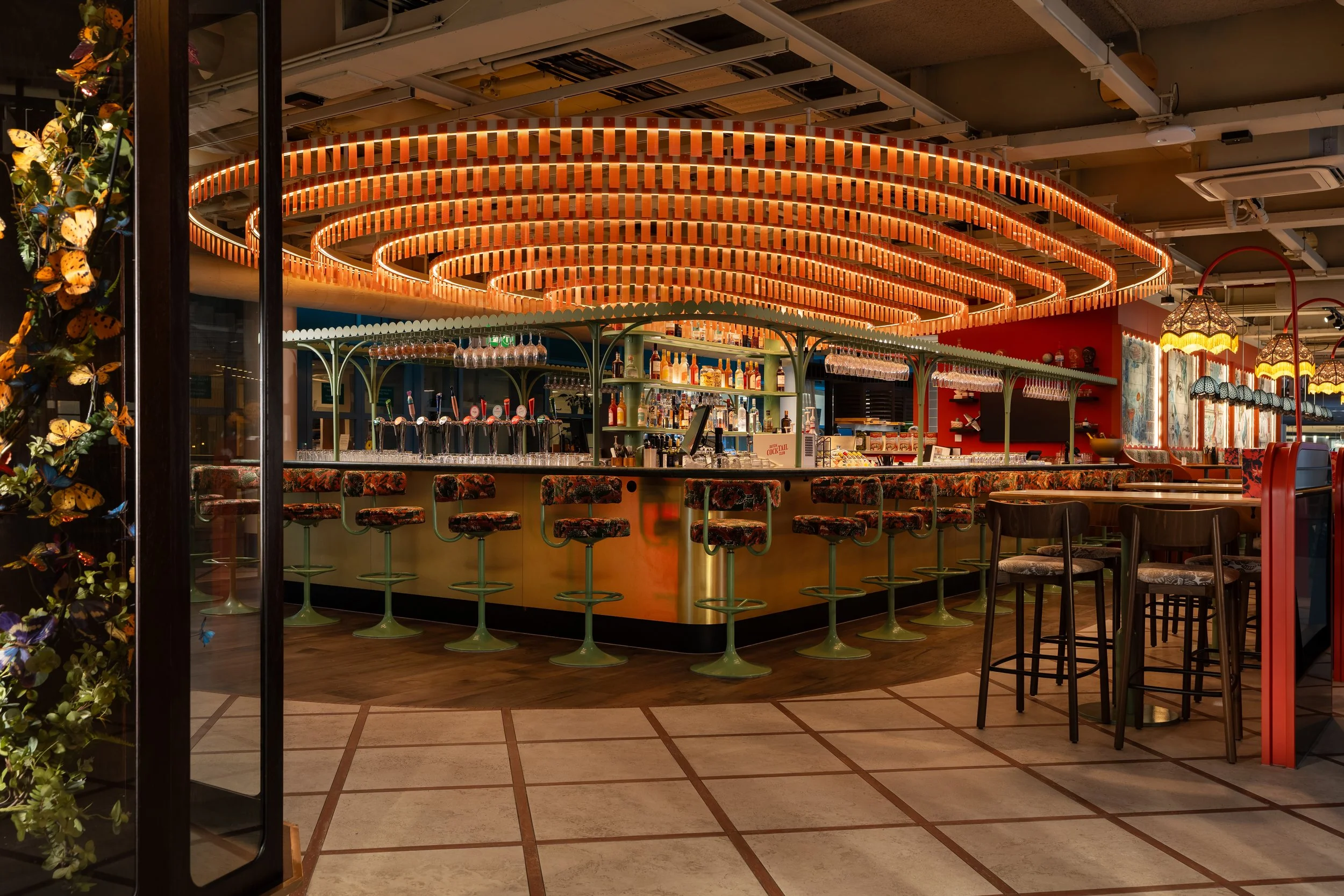 A colorful bar with an illuminated, spiral ceiling feature, bar stools with floral upholstery, and a decorated counter with glasses and bottles.