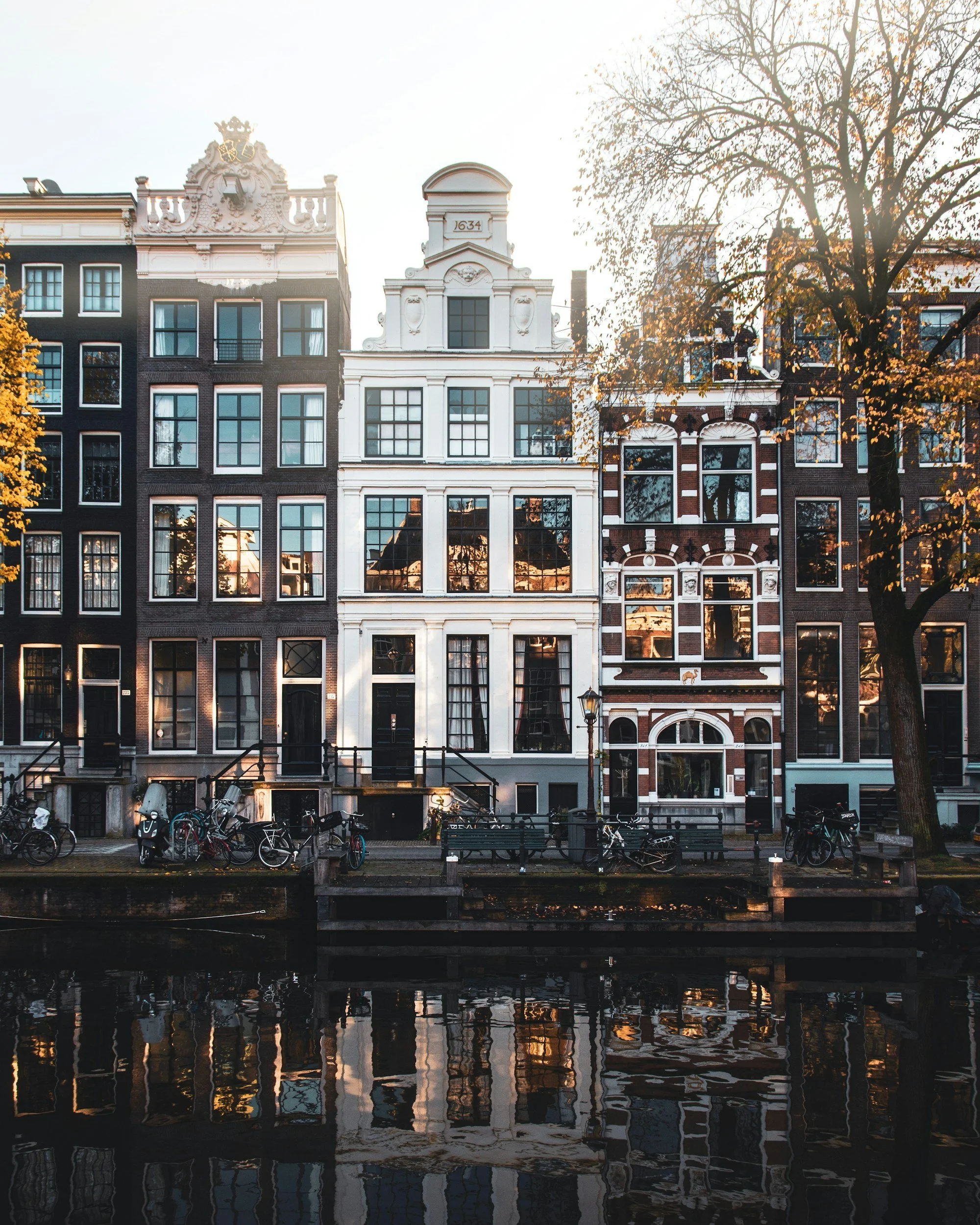 A row of tall, narrow European-style buildings with large windows, situated along a canal with bicycles parked in front, in an urban setting during autumn.