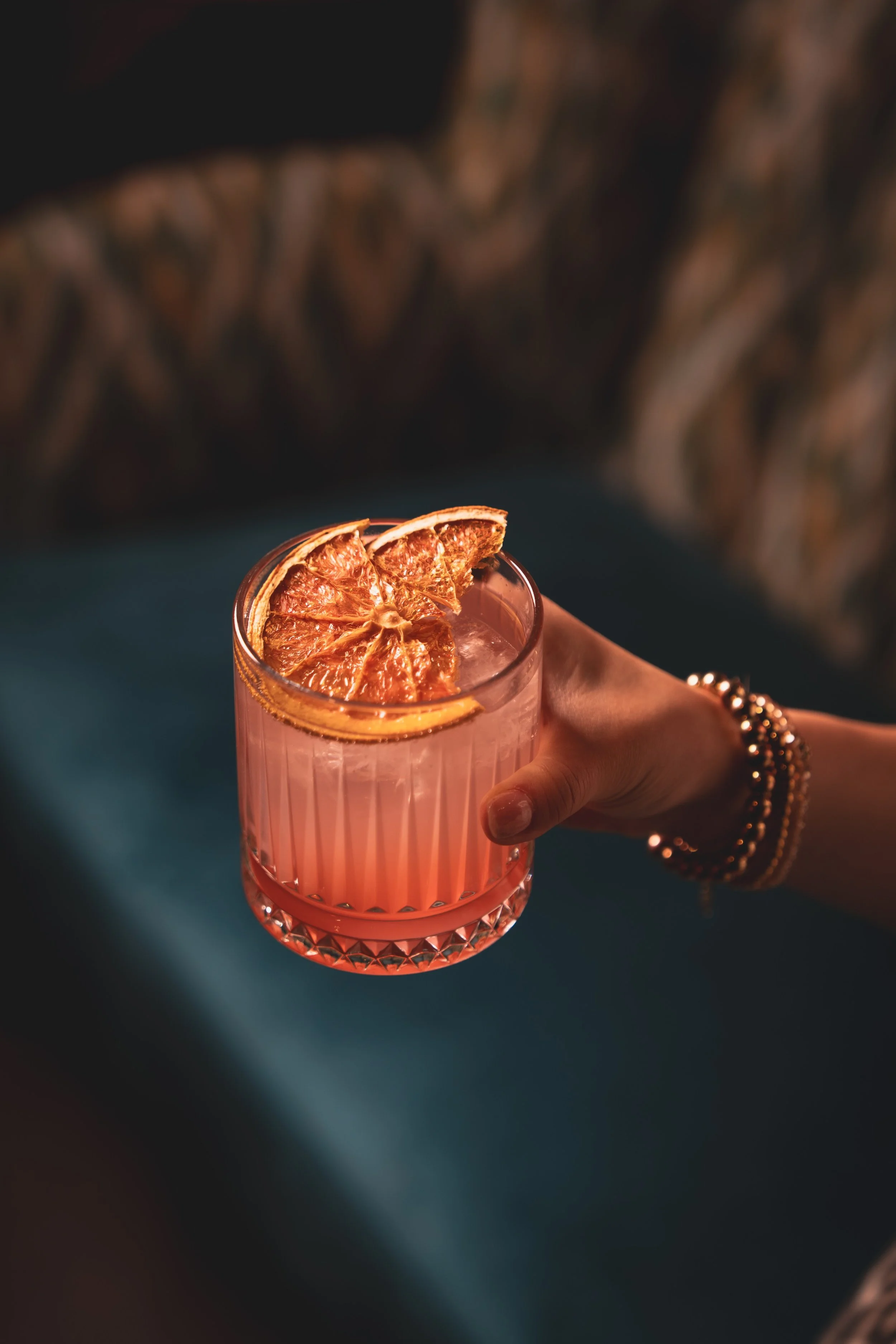A hand holding a glass of pink cocktail garnished with dried orange slices, with a dark, blurred background.