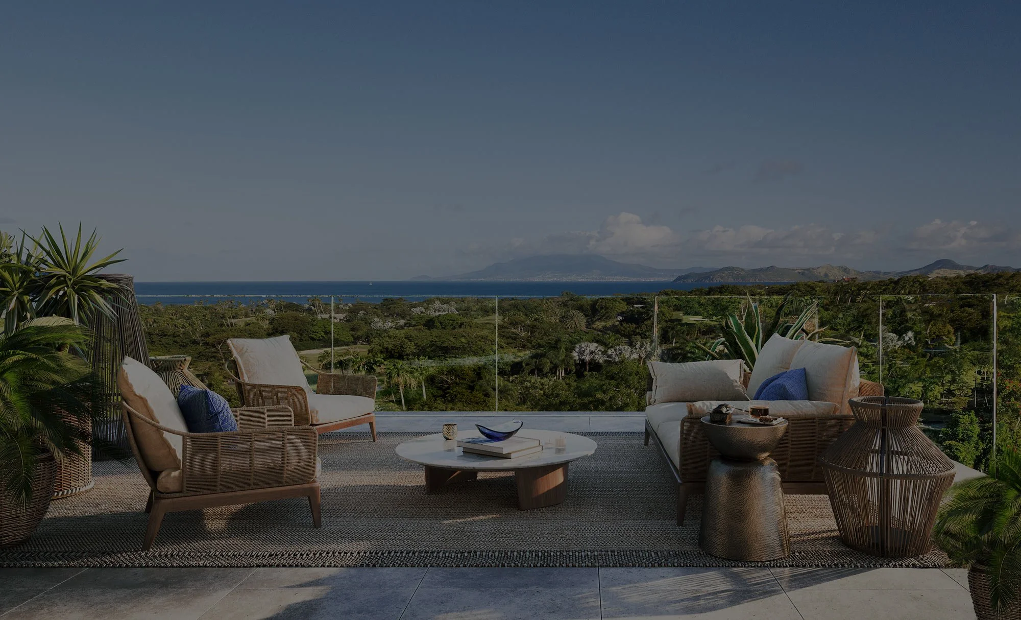 Outdoor balcony with wicker furniture including sofas and chairs with cushions, a white round coffee table, potted plants, and a scenic view of the ocean, green trees, and mountains under a cloudy sky.