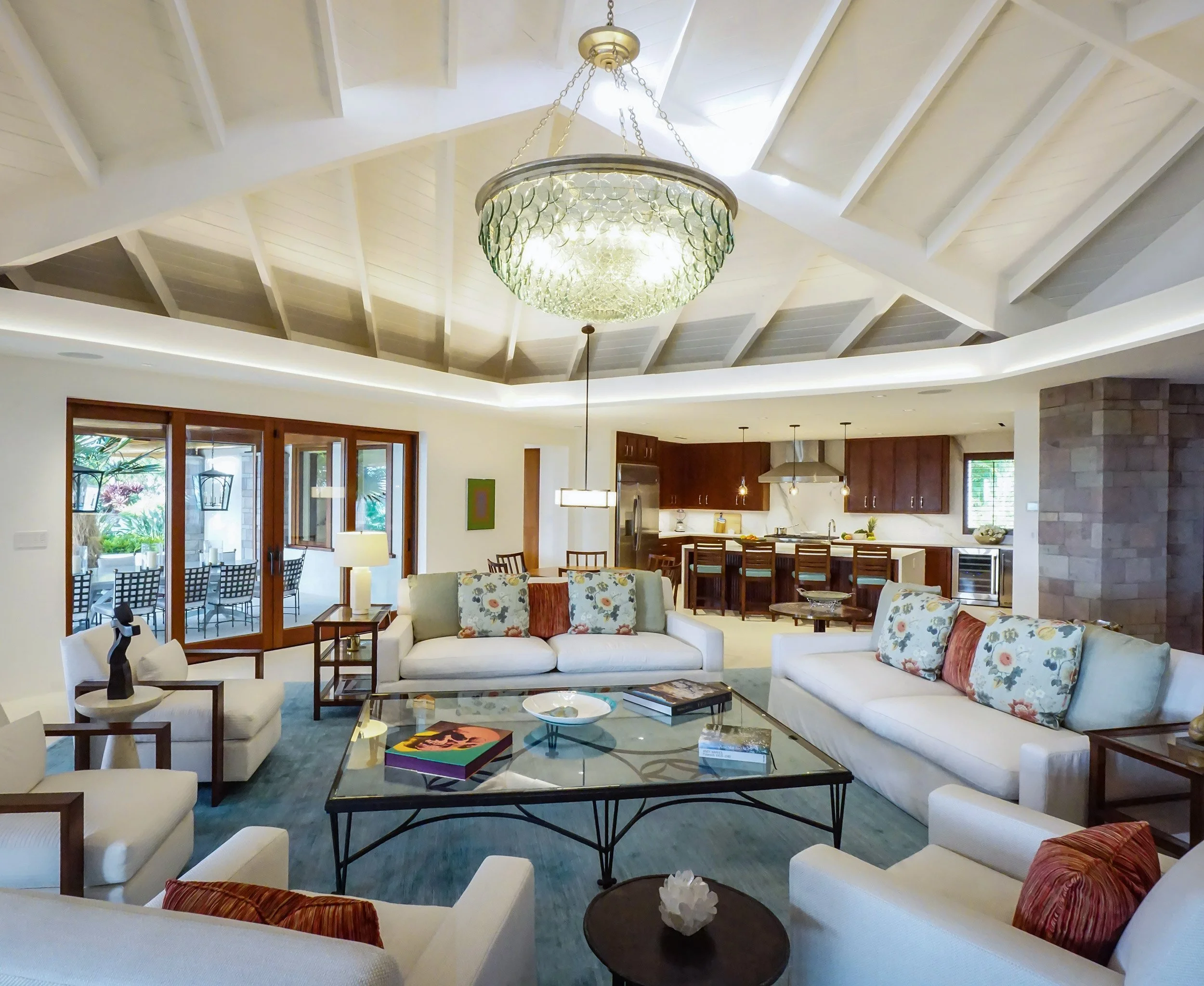 A spacious, modern living room with white sofas and floral accent pillows, a glass coffee table with books, a chandelier hanging from the ceiling, and large windows with wooden frames and sliding doors leading outside.
