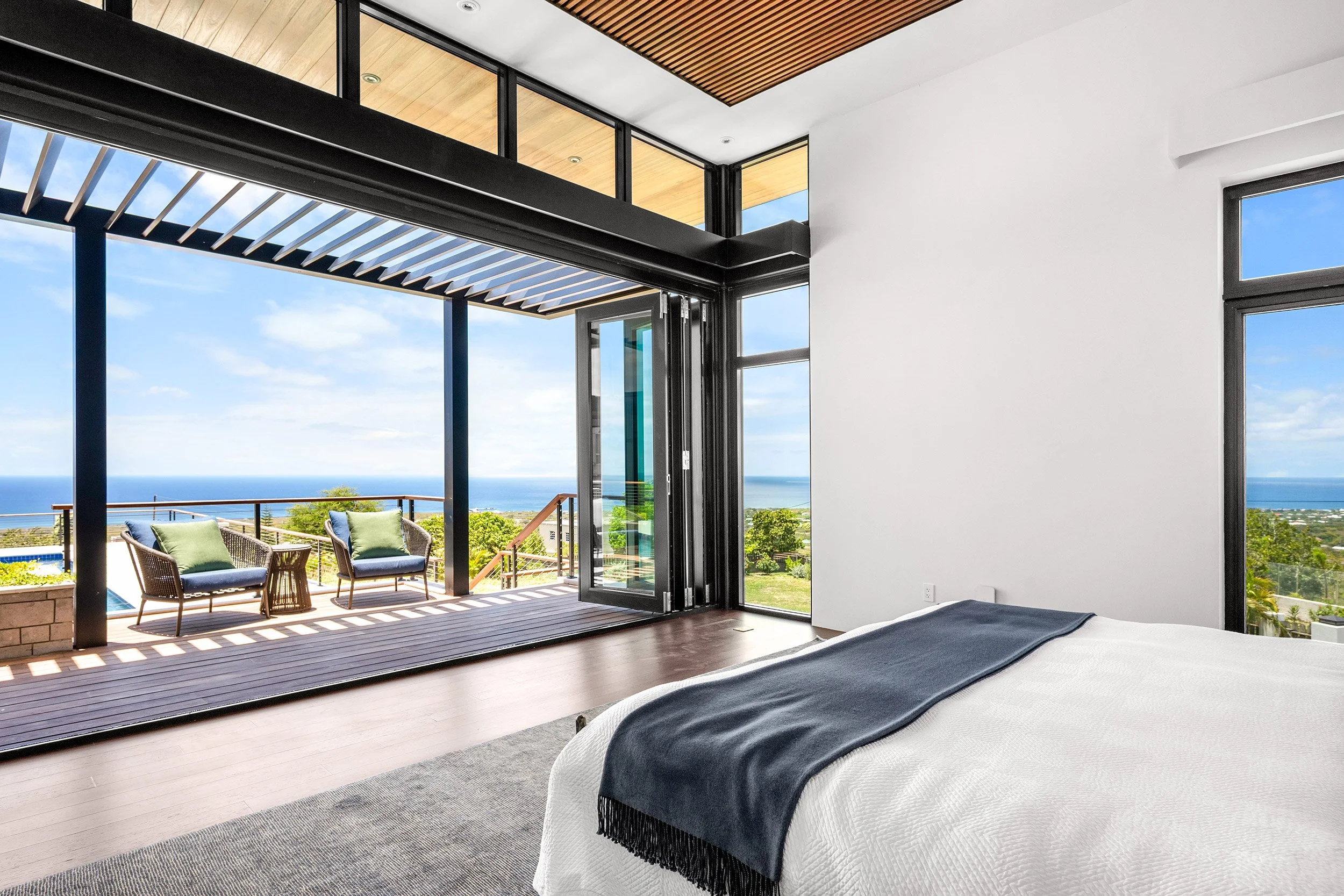 Modern bedroom with sliding glass doors leading to a balcony with outdoor chairs and a view of the ocean, blue sky, and greenery.