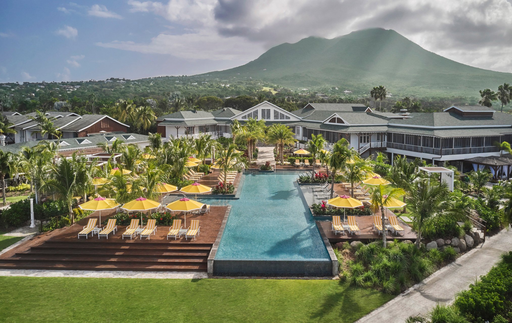 A large outdoor swimming pool surrounded by yellow and white striped lounge chairs and yellow umbrellas, palm trees, and tropical plants, with a large mountain in the background and houses and greenery on the hillside.