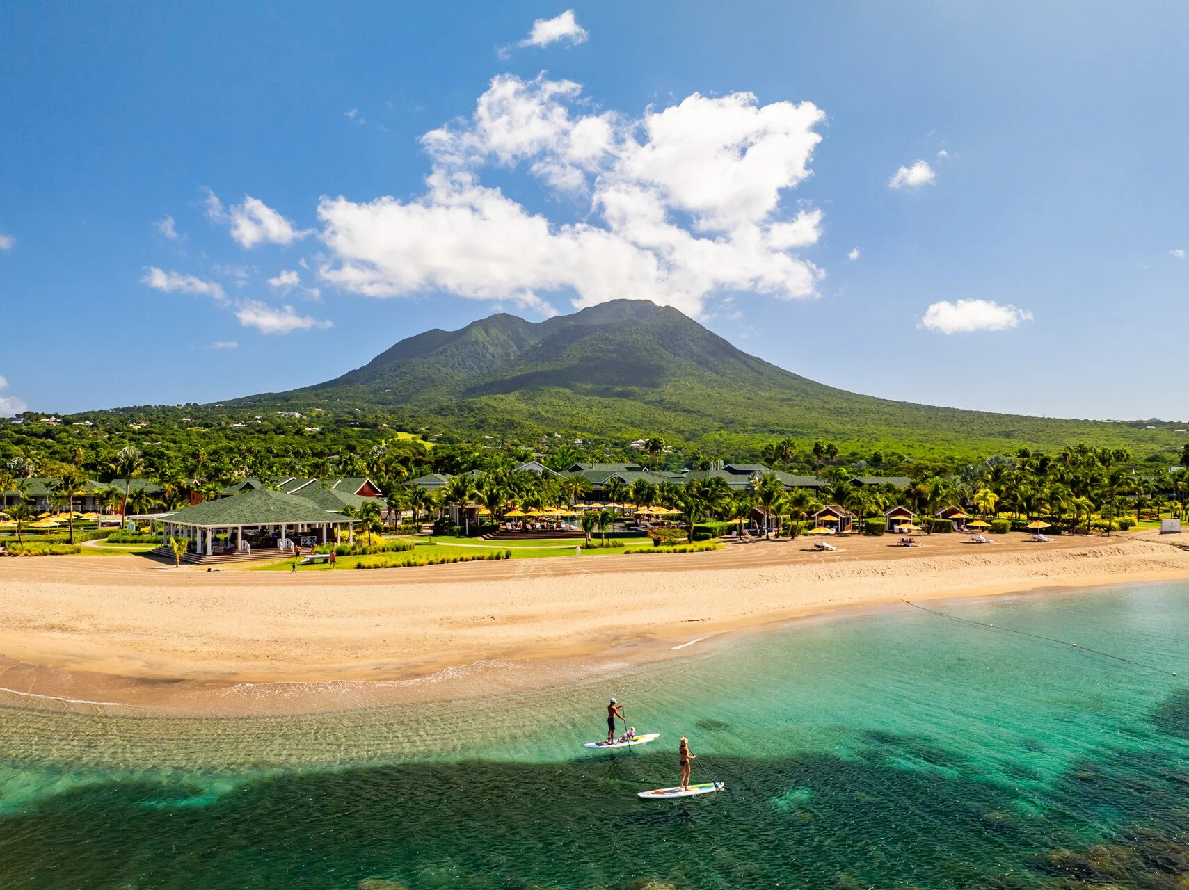 A tropical beach with two people paddleboarding in clear green water, a sandy shoreline with beach chairs, a lush green resort area with palm trees, and a large mountain in the background under a blue sky with scattered clouds.