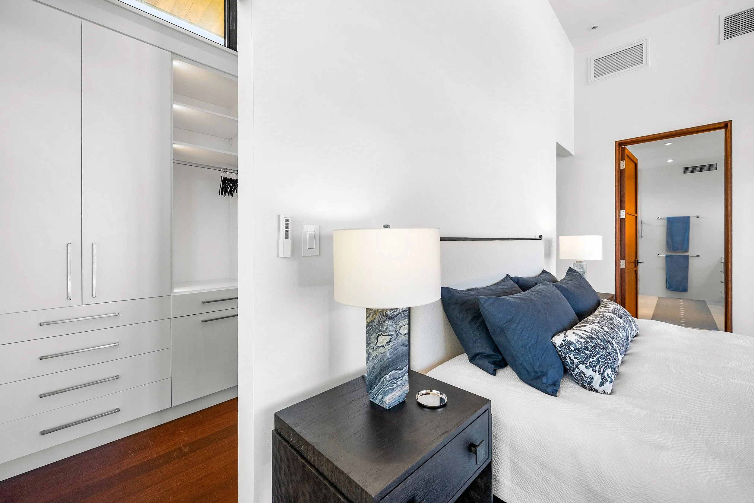 Modern bedroom with white walls, dark hardwood floors, a bed with navy pillows, a nightstand with a lamp, and a doorway with a wooden frame leading to a bathroom.