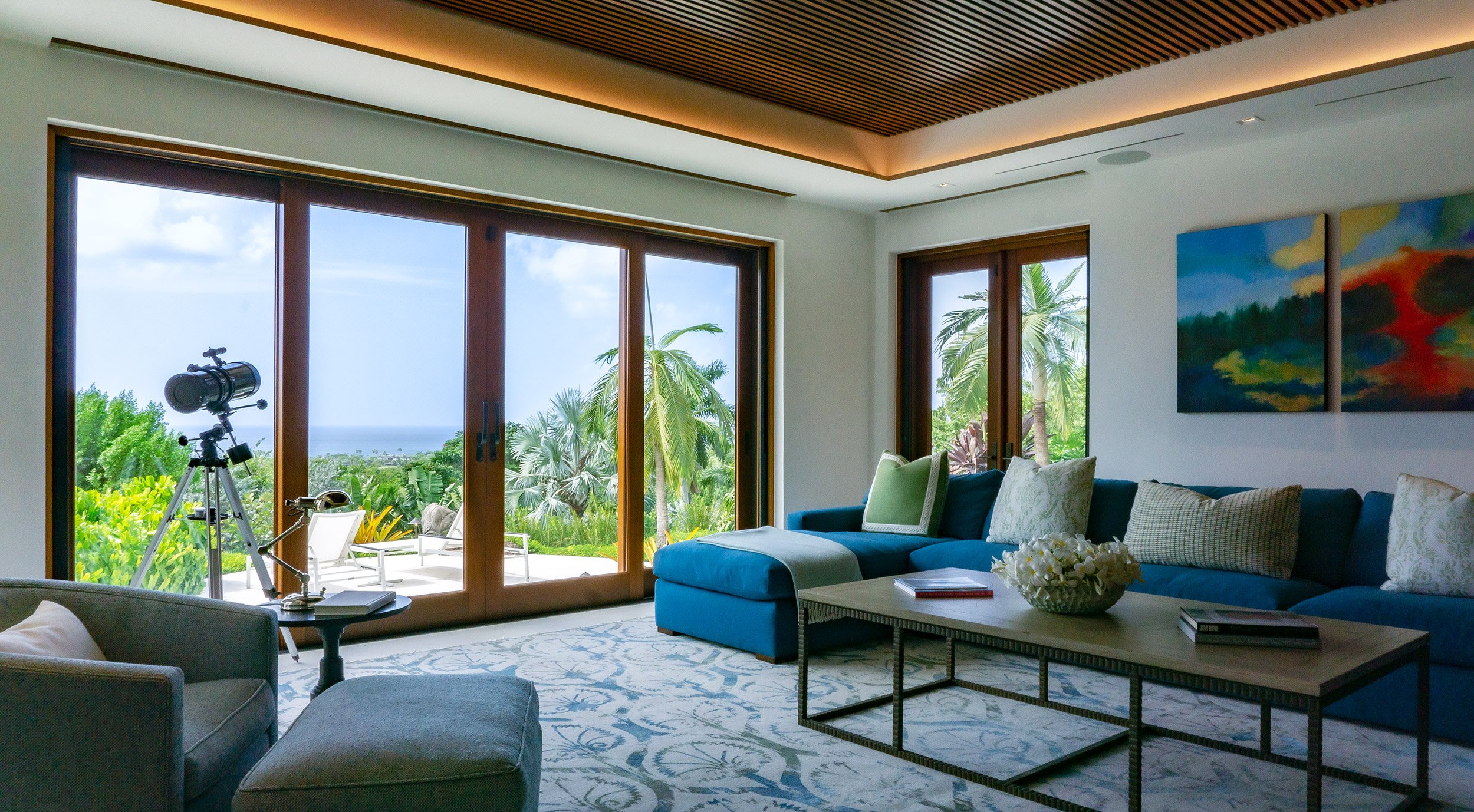 Living room with large glass sliding doors opening to a tropical outdoor view of palm trees and the ocean. There is a telescope set up near the doors, a blue sectional sofa with cushions, a coffee table with a bowl of flowers, and colorful abstract artwork on the wall.
