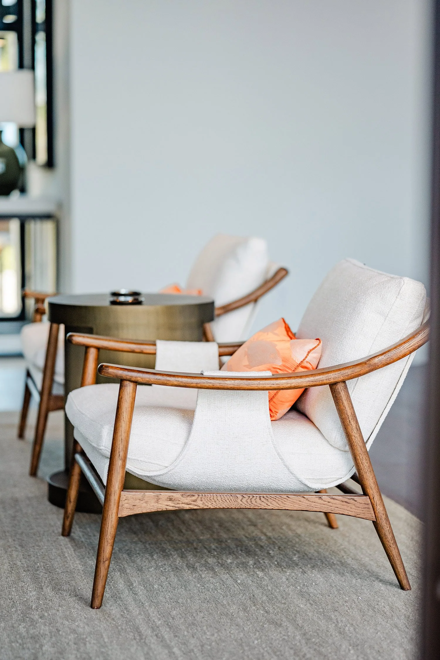 A row of beige upholstered armchairs with wooden arms and legs, each with orange pillows, arranged along a wall in a living room. A black side table is in front of the first chair.