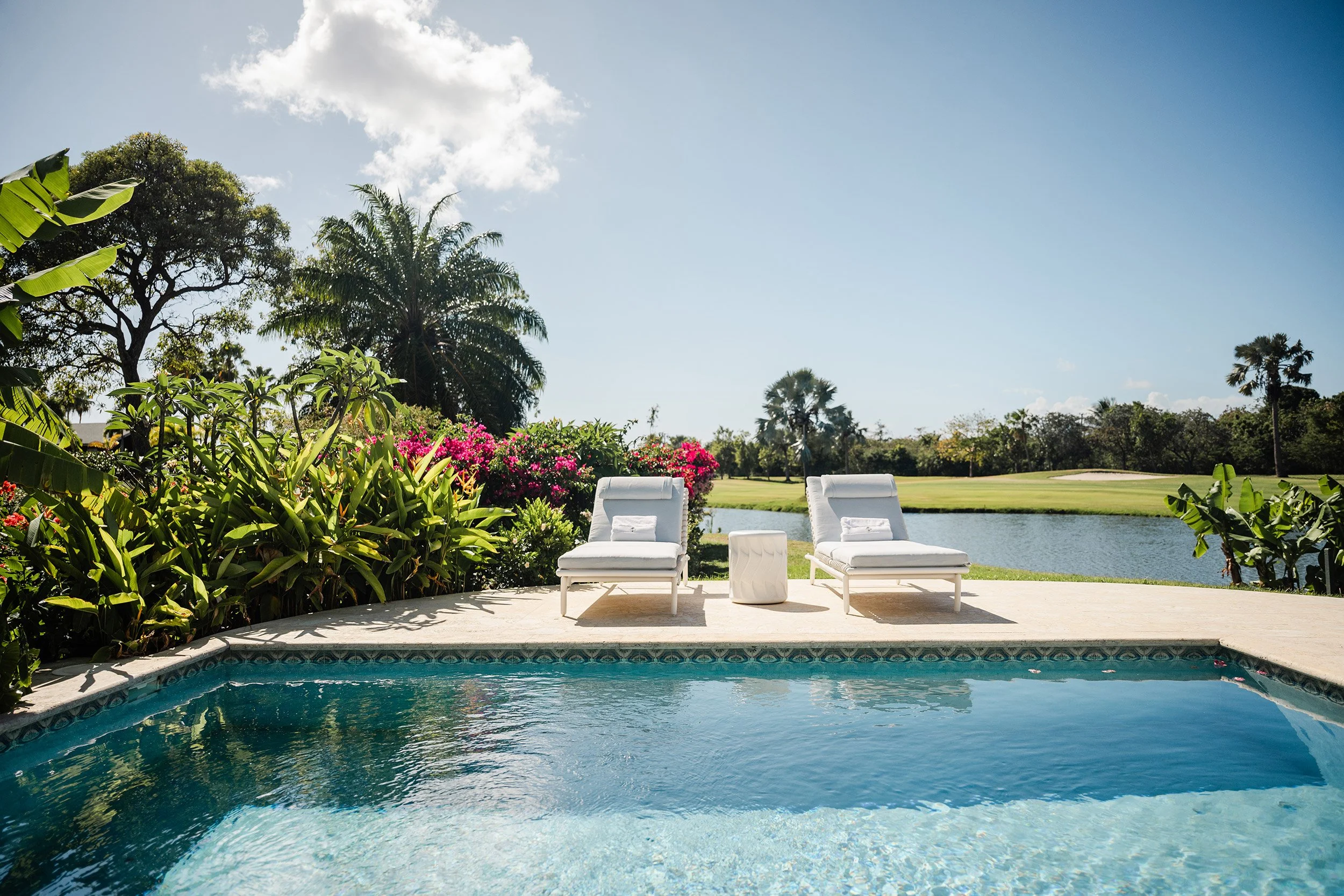 Poolside view with two white lounge chairs, a small white table, lush green plants, trees, a pond, and a clear blue sky with some clouds.