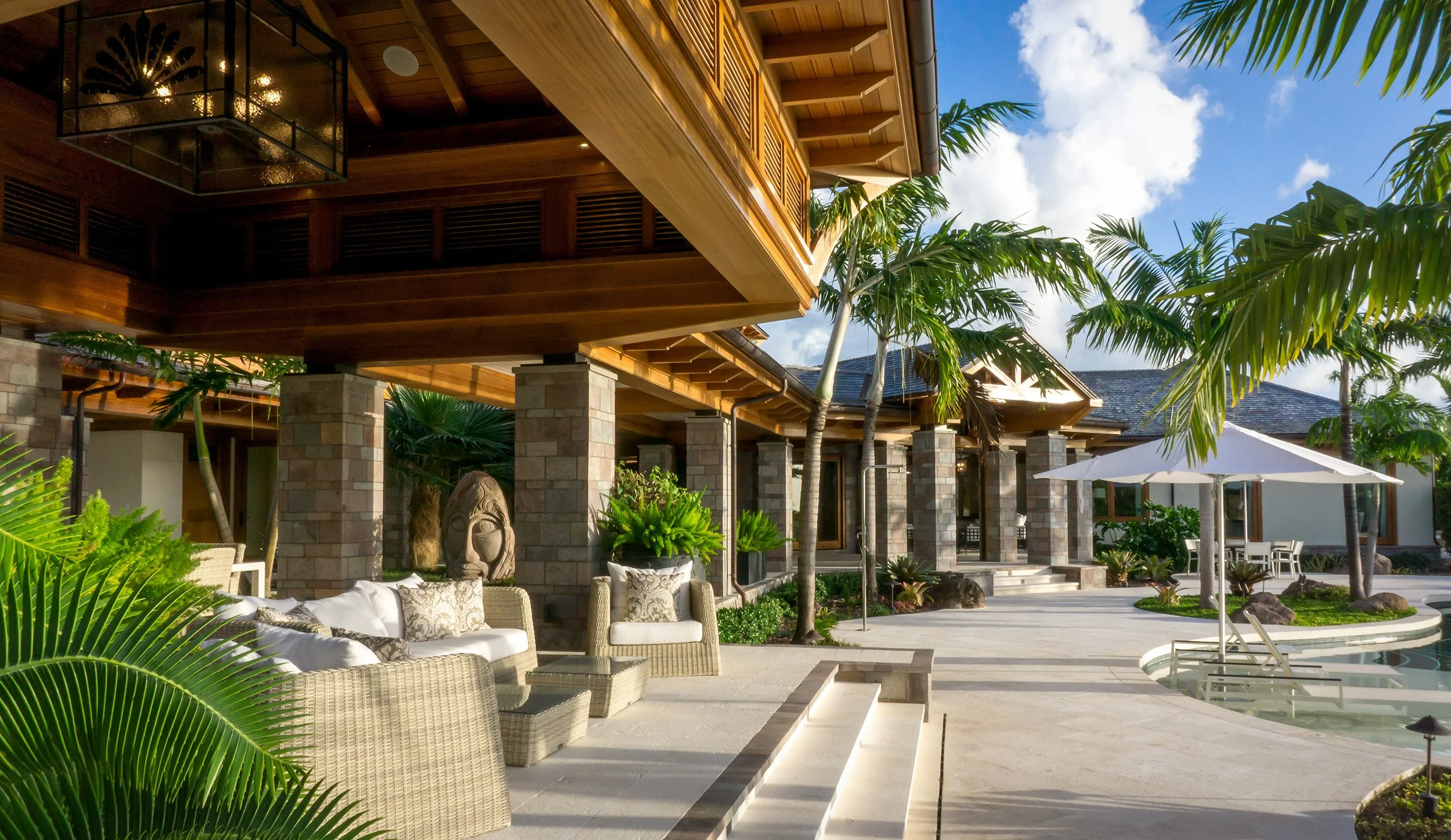 Luxury house with a pool, surrounded by tropical plants, wicker furniture with cushions, and a white umbrella in the backyard, under a partly cloudy sky.
