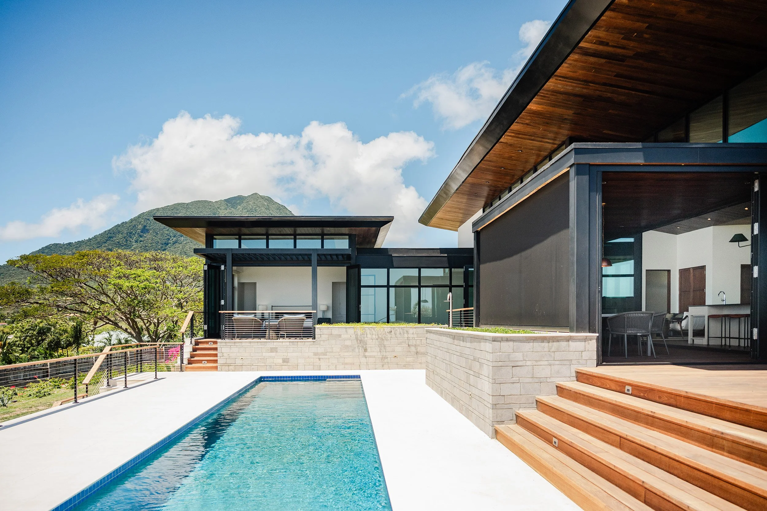 Modern house with wooden accents and large glass windows, overlooking a mountain with a pool in the foreground, outdoor seating area, and steps leading to the pool, under a blue sky with scattered clouds.