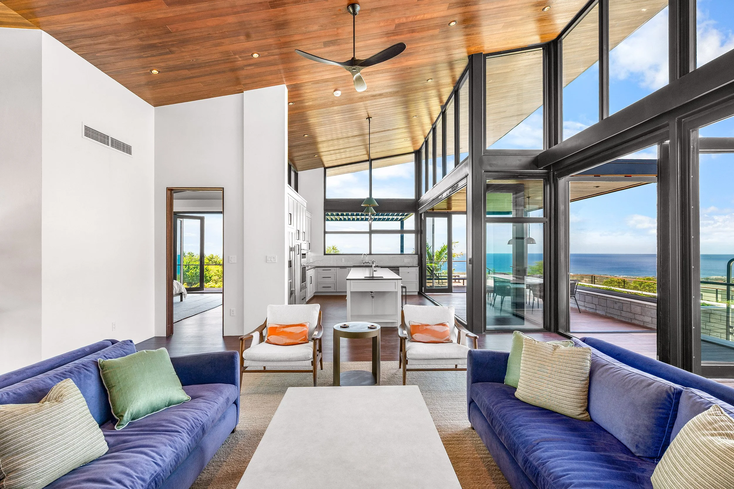 Living room with two blue sofas, cream armchairs with orange pillows, and a white coffee table, overlooking a balcony with a view of the ocean through large windows.