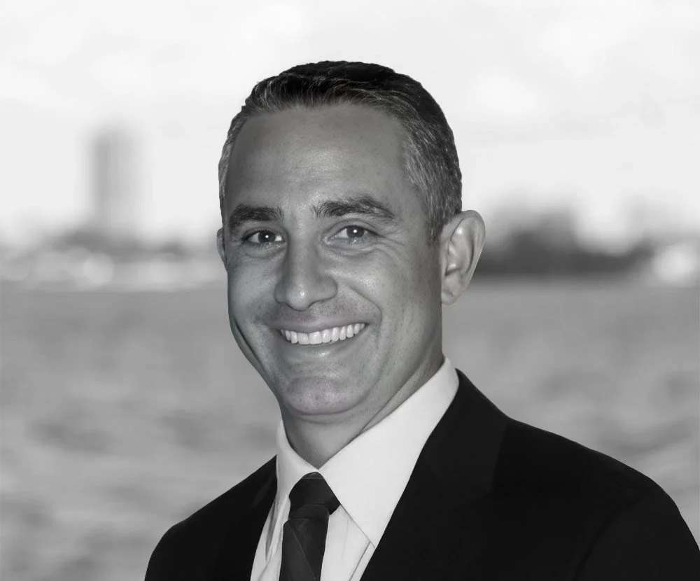 Black and white portrait of a smiling man in a suit, background showing a waterfront with blurred cityscape.