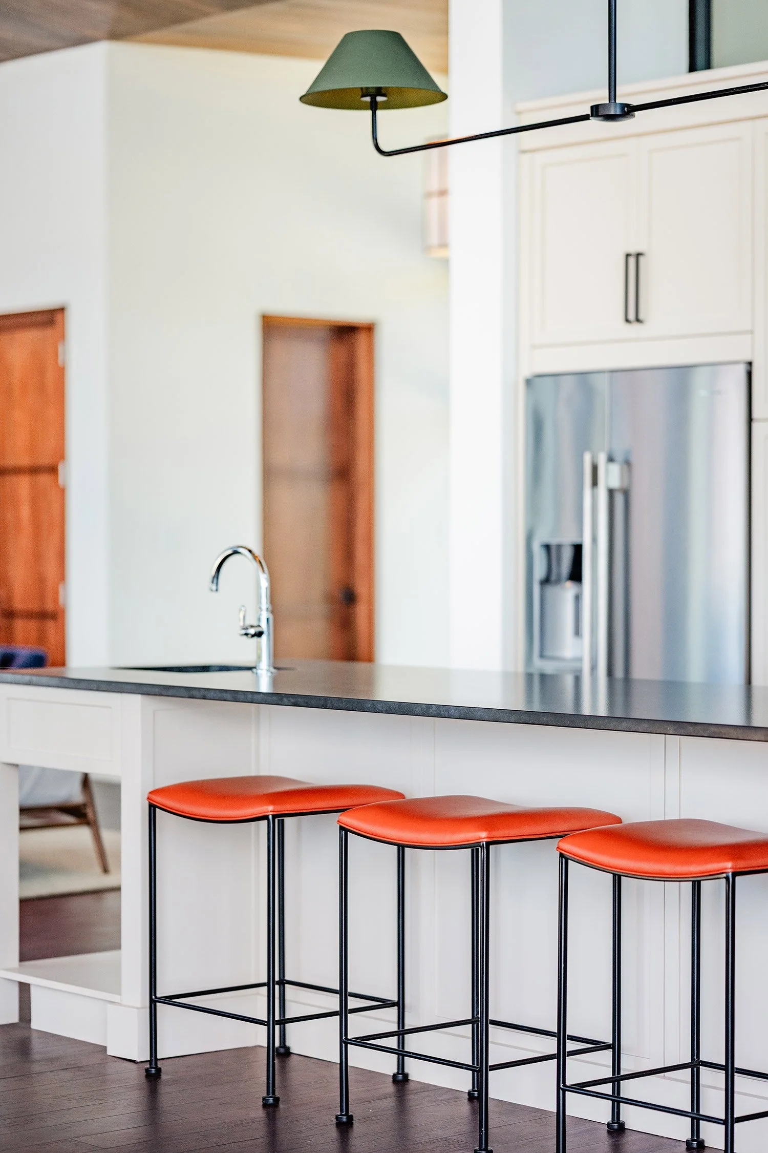 Modern kitchen with white island counter, black countertops, three bar stools with red cushions and black metal legs, stainless steel refrigerator, and light-colored cabinets.