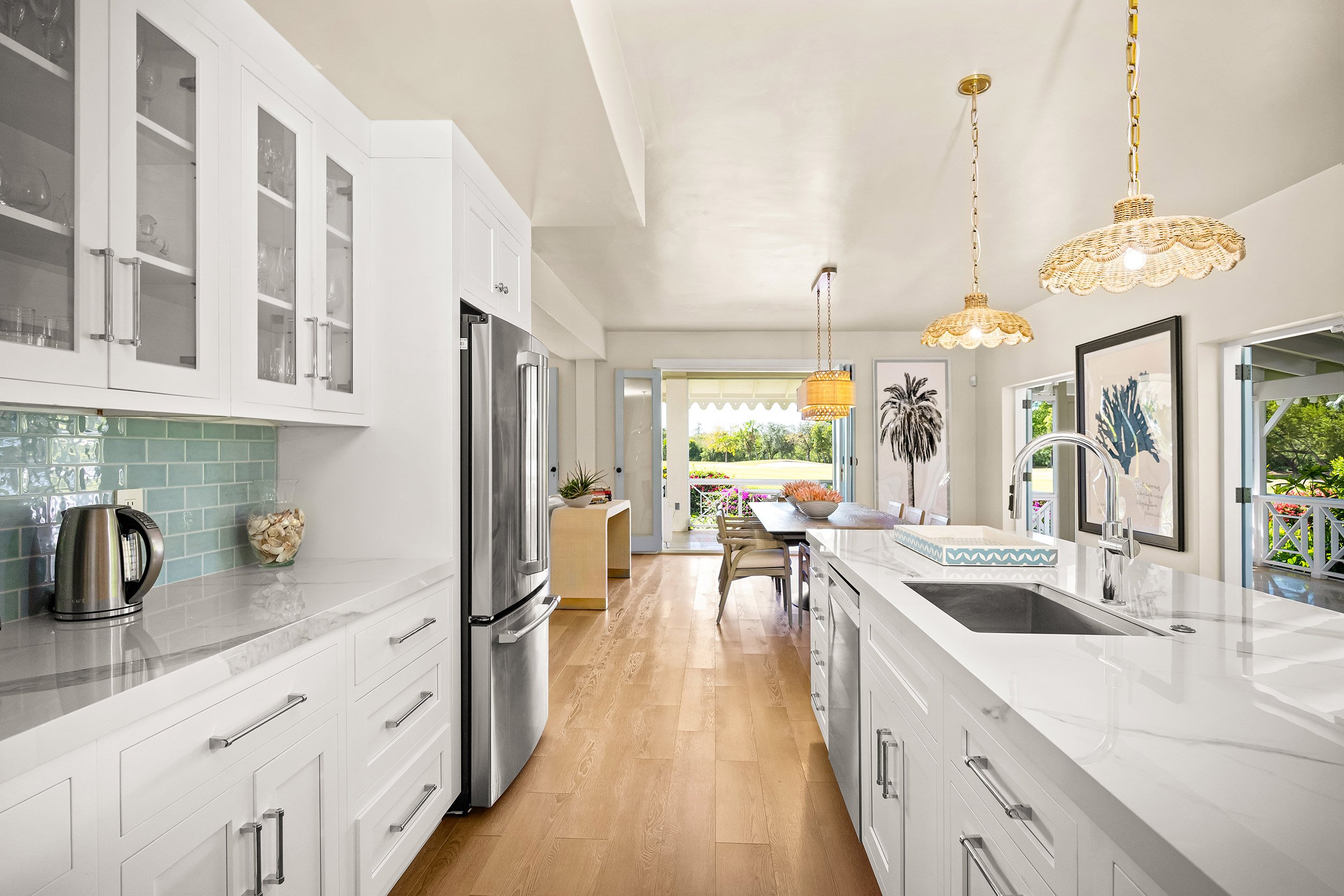 Bright modern kitchen with white cabinets, stainless steel appliances, wooden flooring, and large windows opening onto a garden patio with chairs and colorful flowers.