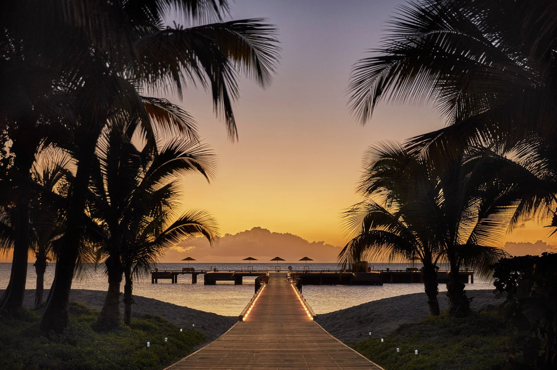 Sunset over a tropical beach seen from a wooden pier lined with small lights, flanked by palm trees with fronds silhouetted against the sky, with umbrellas and chairs on the beach.