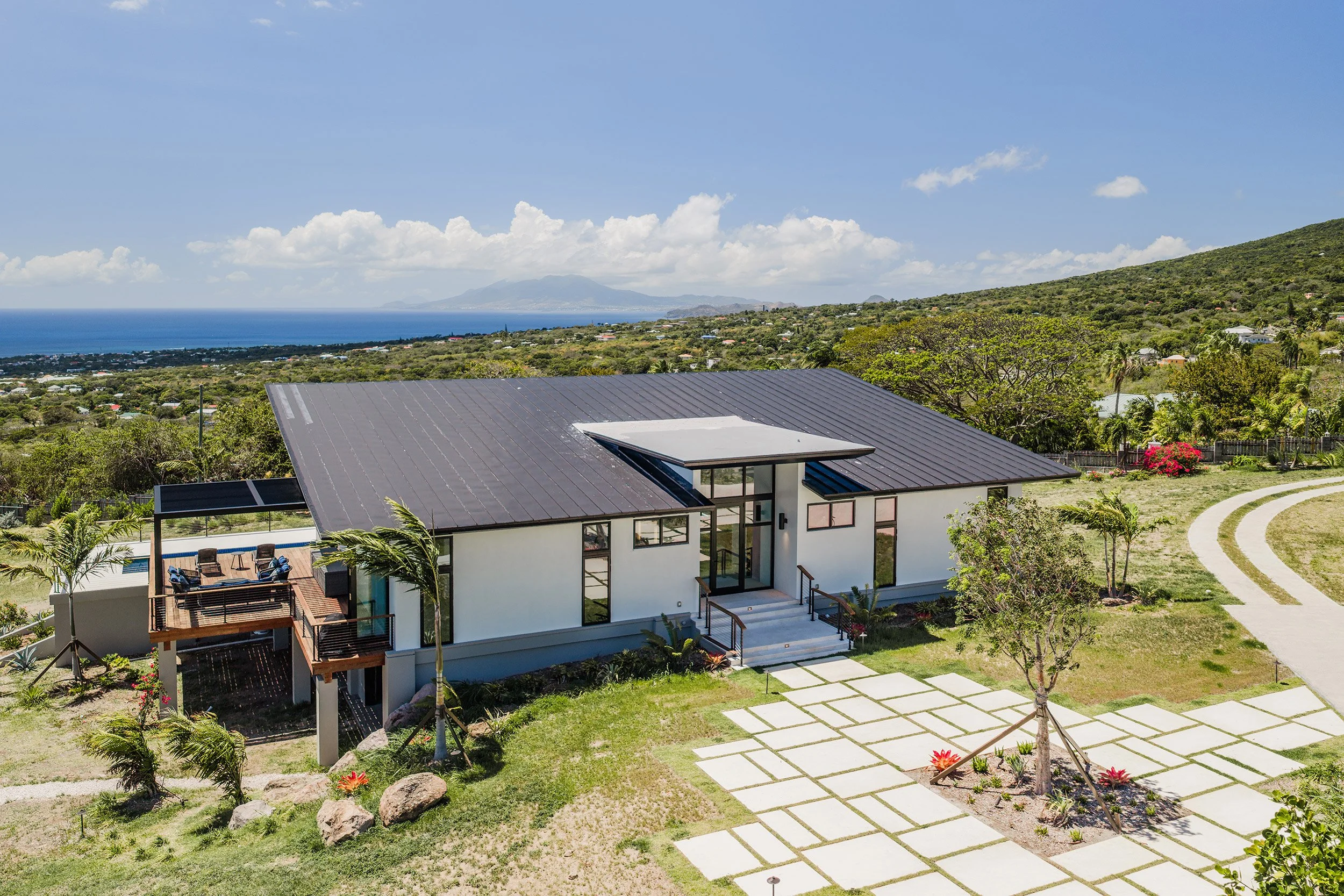 A modern house with a black metal roof and large glass windows in a lush, green landscape with trees, a winding pathway, and mountain and ocean views in the background.