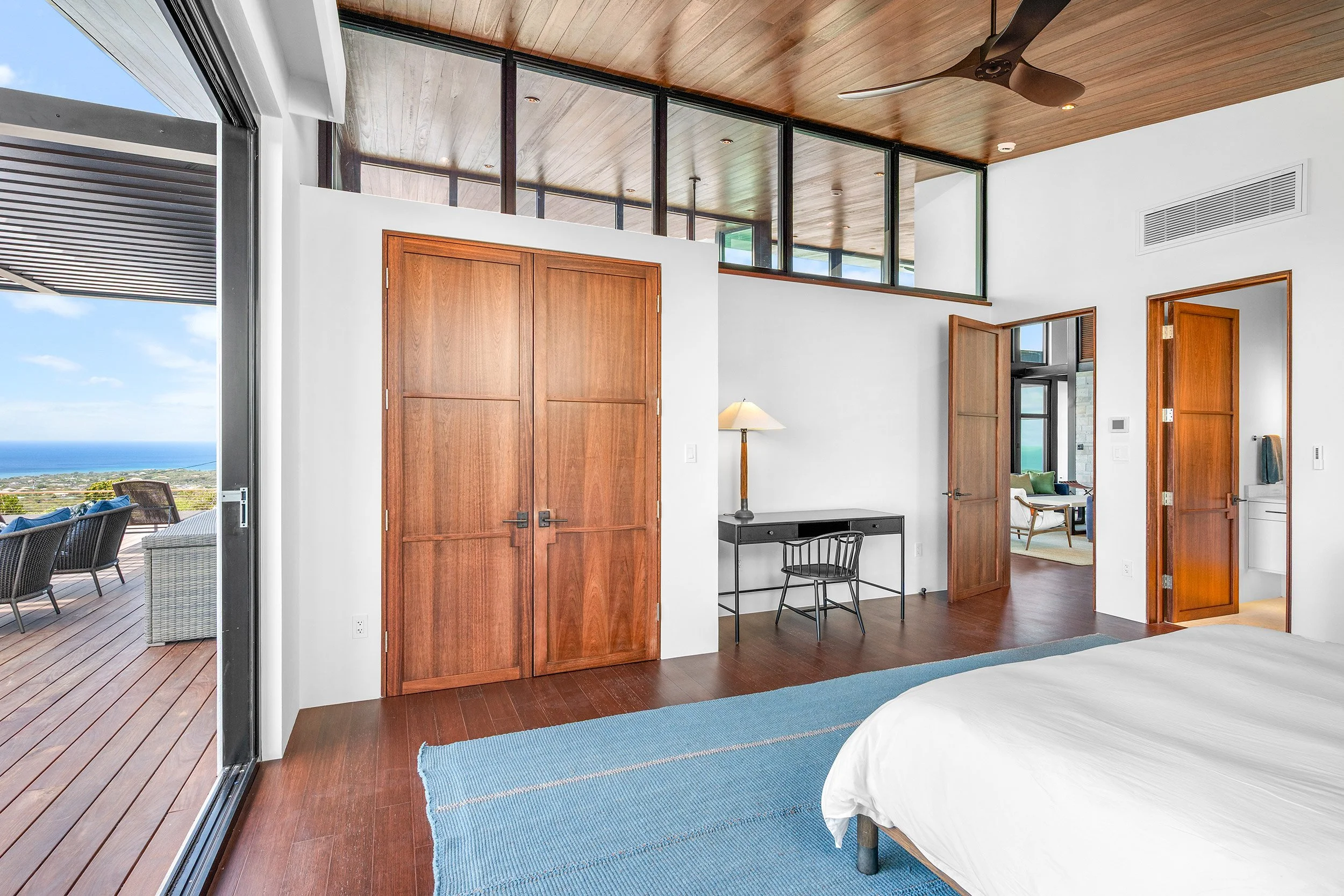 Modern bedroom with a sliding glass door opening to an outdoor deck with seating, overlooking a scenic view. The room features wooden doors, a blue rug, a black desk with a chair, and a ceiling fan.
