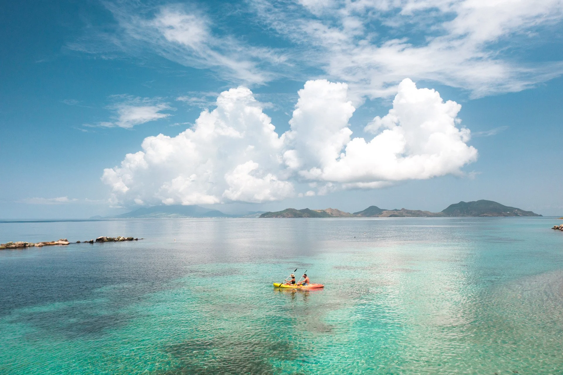 Two people kayaking in turquoise waters off a rocky coastline, with distant mountains and a partly cloudy sky.